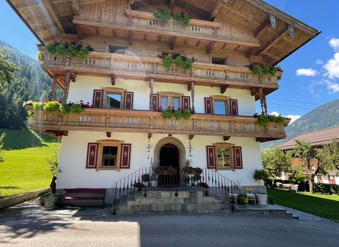 Mehrstöckiges Haus im alpinen Stil mit Holzbalconien, Blumen und grüner Umgebung bei sonnigem Himmel.