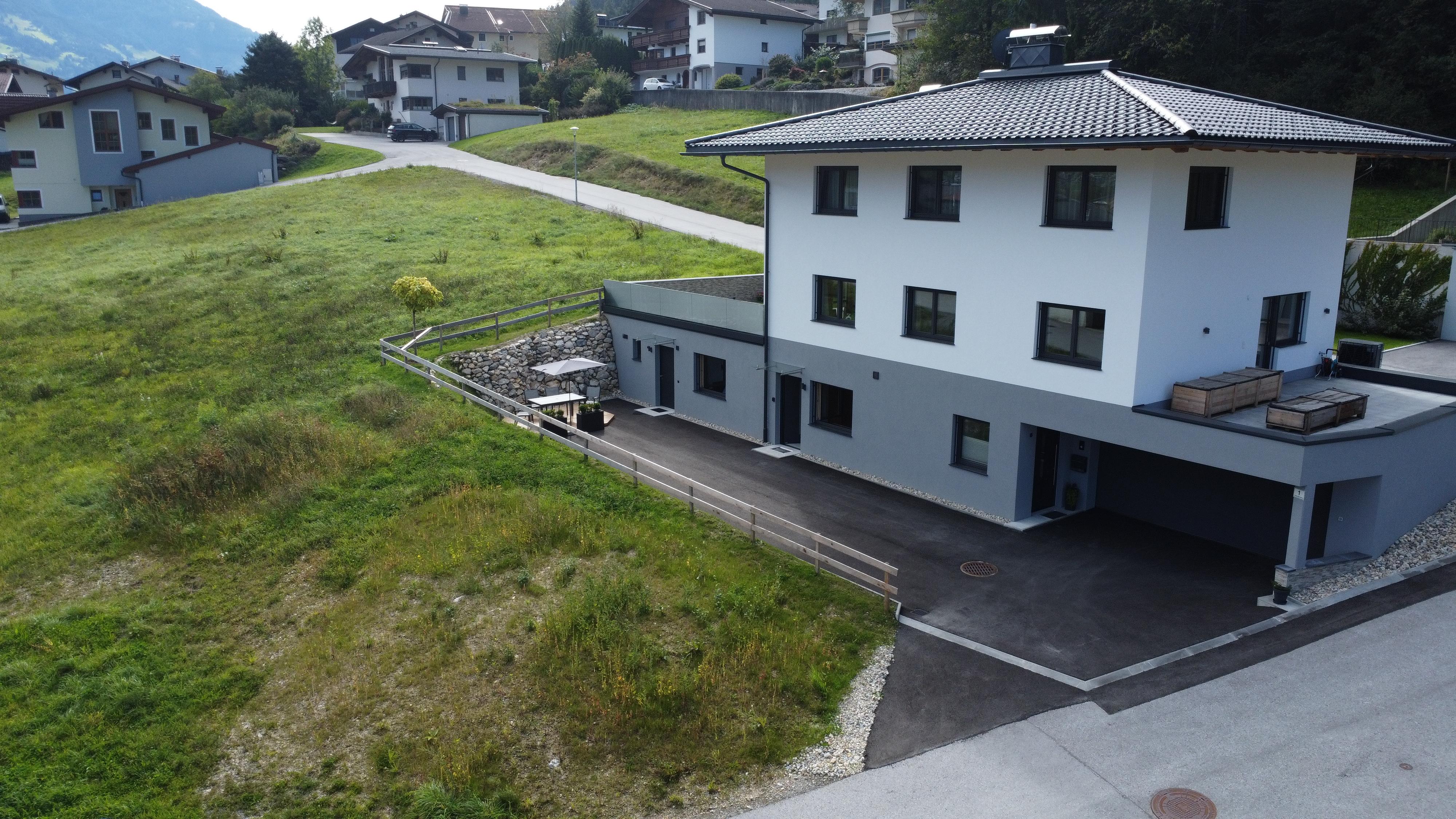 Modern white house with terrace, surrounded by green hills and neighboring houses in a mountainous area.
