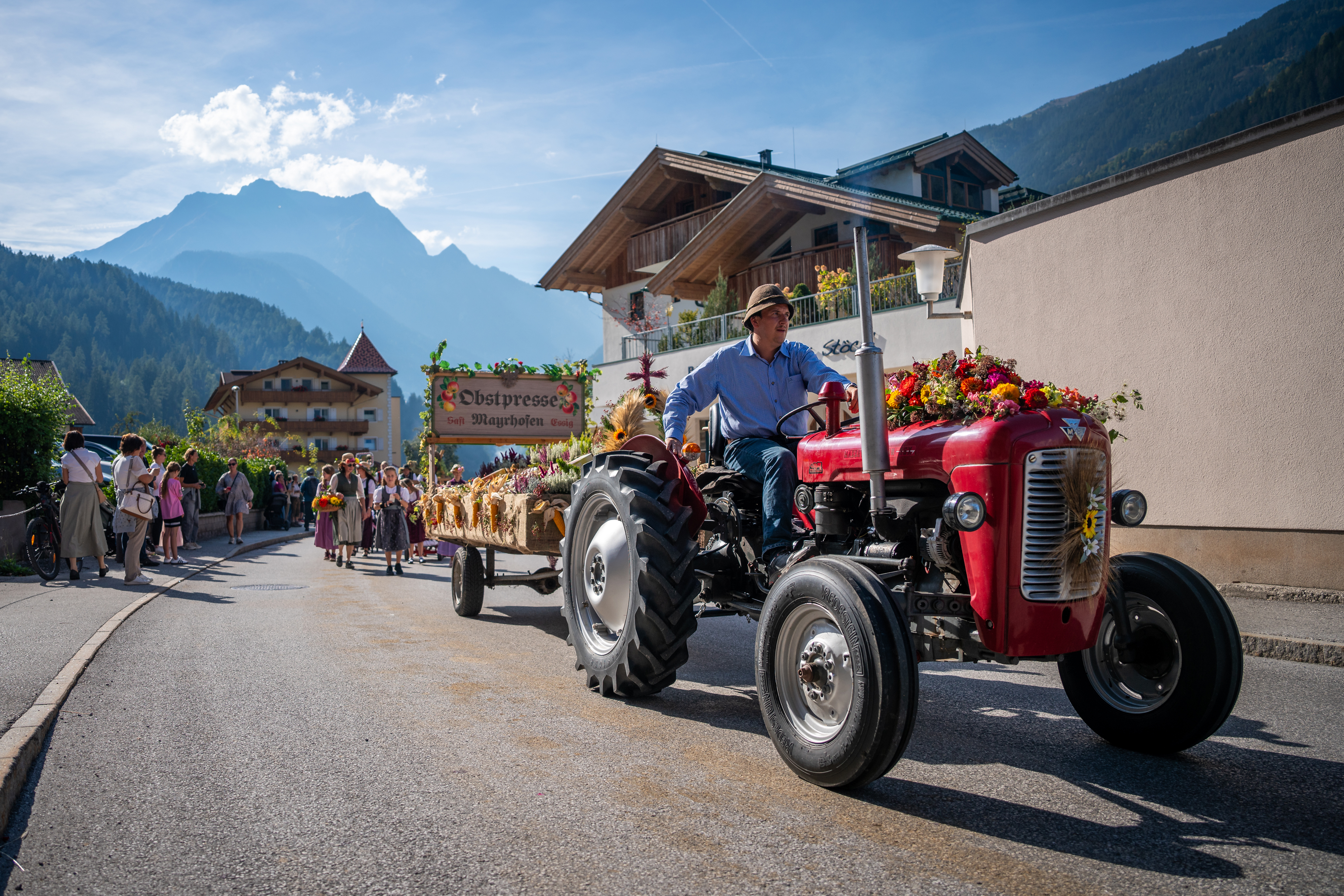 Ein Festzug beim Zillertaler Bauern- und Musikherbst: roter Oldtimer-Traktor zieht einen geschmückten Holz-Festwagen mit Blumenschmuck über die Dorfstraße, links Zuschauer, dahinter schroffe Berge unter blauem Himmel.