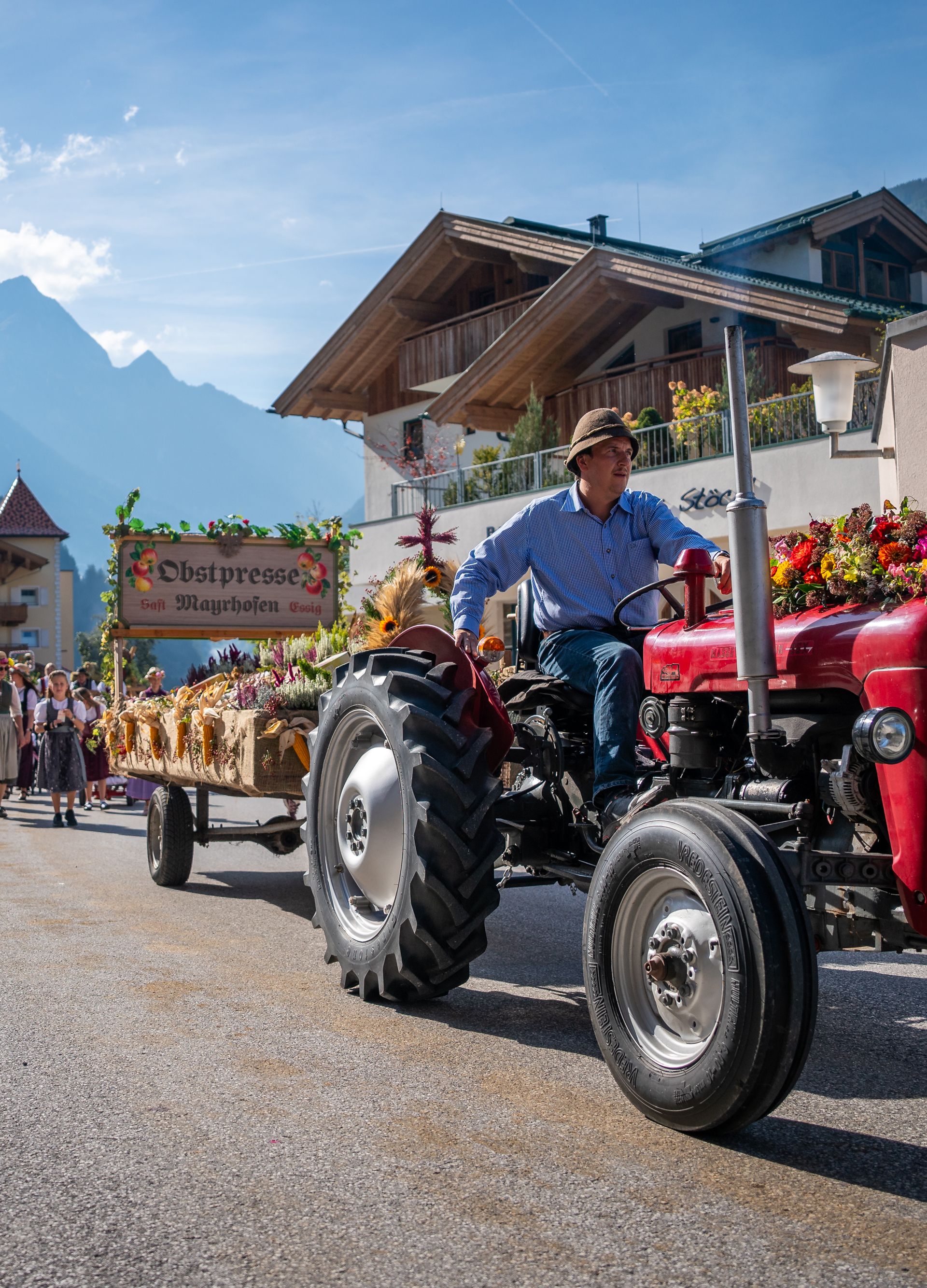 Ein Festzug beim Zillertaler Bauern- und Musikherbst: roter Oldtimer-Traktor zieht einen geschmückten Holz-Festwagen mit Blumenschmuck über die Dorfstraße, links Zuschauer, dahinter schroffe Berge unter blauem Himmel.