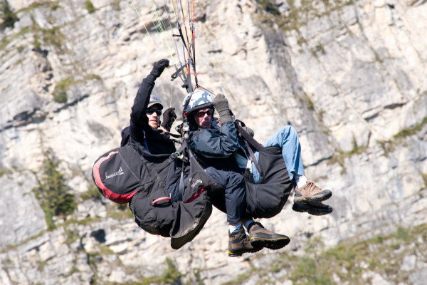 Zwei Paraglider fliegen über eine felsige Berglandschaft, beide tragen Schutzkleidung und Sonnenbrillen.