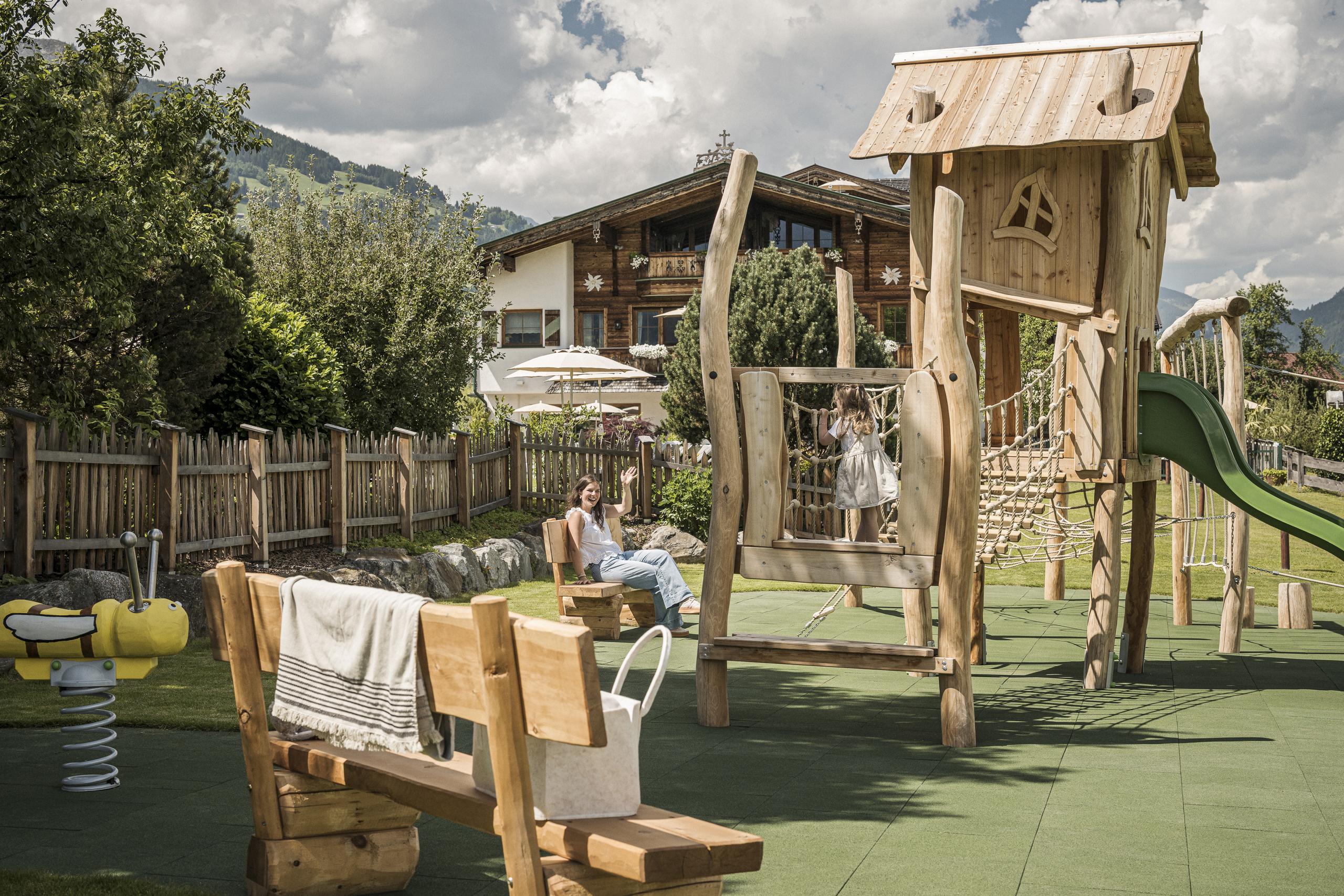 Children playing on wooden playground equipment in a garden with trees and a house behind.