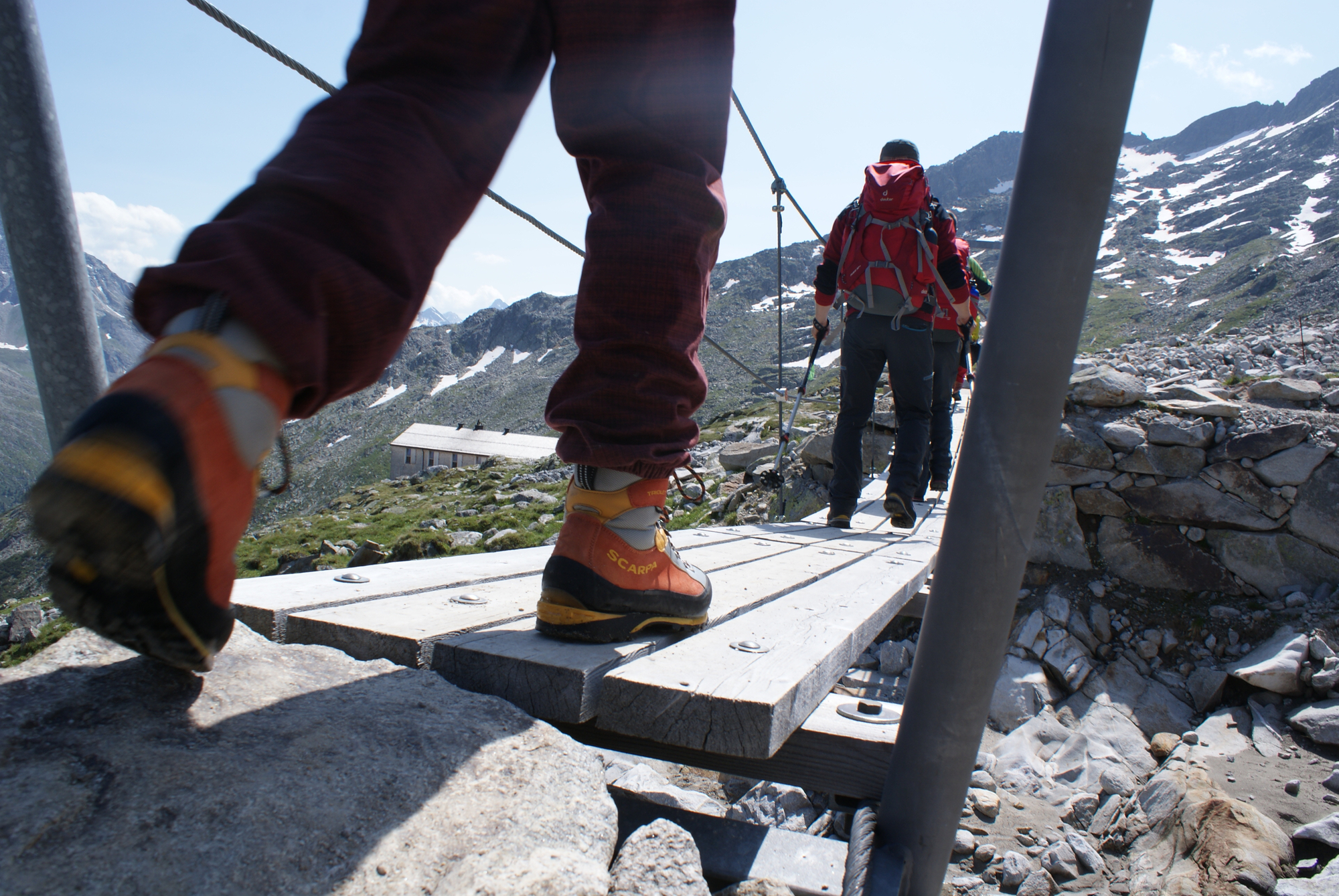 Hikers on the Peter Habeler Trail cross a narrow wooden bridge in high alpine terrain. With backpacks and hiking boots, they make their way through rocky terrain, surrounded by snow-capped peaks and clear mountain air.