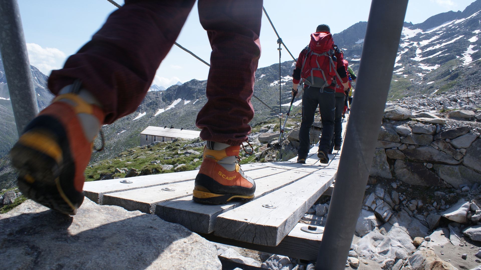 Wanderer auf der Peter-Habeler-Runde überqueren eine schmale Holzbrücke in hochalpiner Landschaft. Mit Rucksäcken und Bergschuhen geht es durch felsiges Gelände, umgeben von schneebedeckten Gipfeln und klarer Bergluft.
