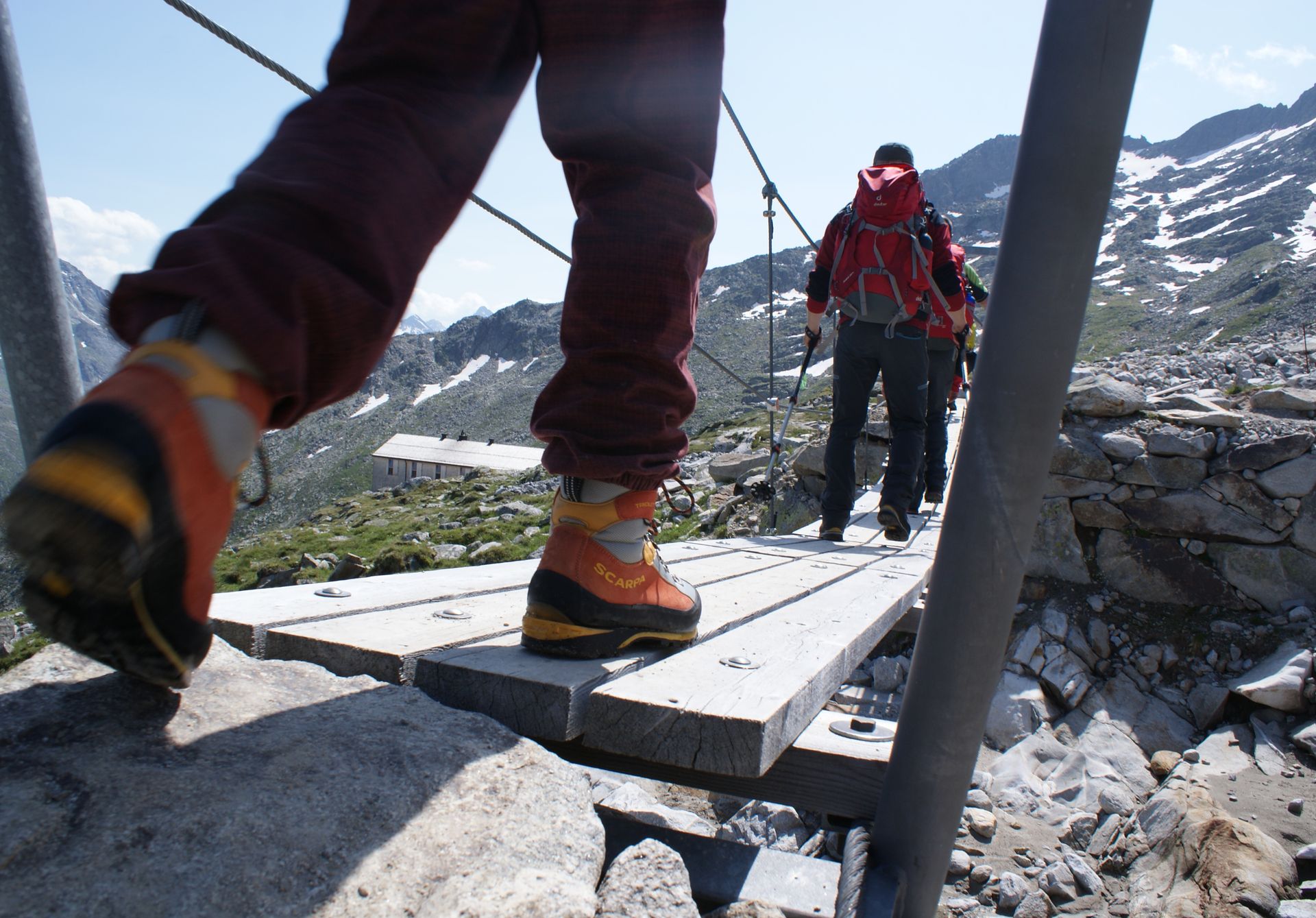 Wanderer auf der Peter-Habeler-Runde überqueren eine schmale Holzbrücke in hochalpiner Landschaft. Mit Rucksäcken und Bergschuhen geht es durch felsiges Gelände, umgeben von schneebedeckten Gipfeln und klarer Bergluft.