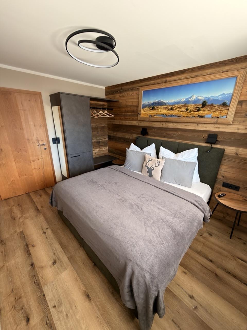 Schlafzimmer mit Holzwand, Bett mit grüner Kopfstütze, Wandbild der Berge, Nachttisch und moderne Deckenleuchte.
