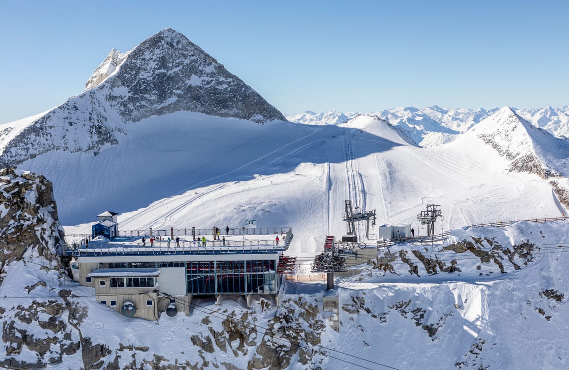 Panorama Terrace Hintertuxer Glacier – Viewing Terrace with Mountain Panorama ©TVB Tux Finkenberg The panorama terrace at the Hintertuxer Glacier: modern building with glass facade and flat roof on a rocky, snow-covered ridge, with steep snow-capped peaks behind under a brilliant blue sky in sunshine.