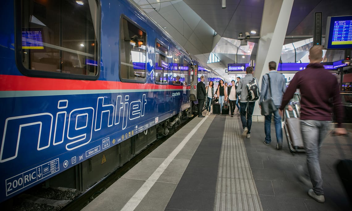 ÖBB - Travel by Nightjet ©ÖBB - Harald Eisenberger ÖBB Nightjet sleeping car on the platform of a modern train station. The blue train with white Nightjet lettering and red stripes is ready to depart. Passengers with luggage move along the illuminated platform.