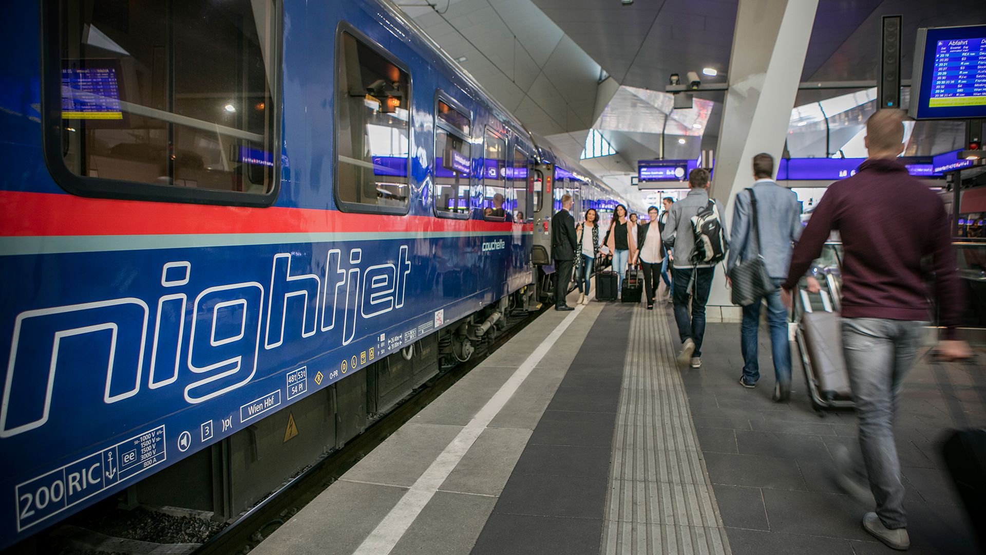 ÖBB Nightjet Schlafwagen am Bahnsteig eines modernen Bahnhofs. Der blaue Zug mit weißer Nightjet-Beschriftung und rotem Streifen steht bereit. Fahrgäste mit Gepäck bewegen sich auf dem beleuchteten Bahnsteig.