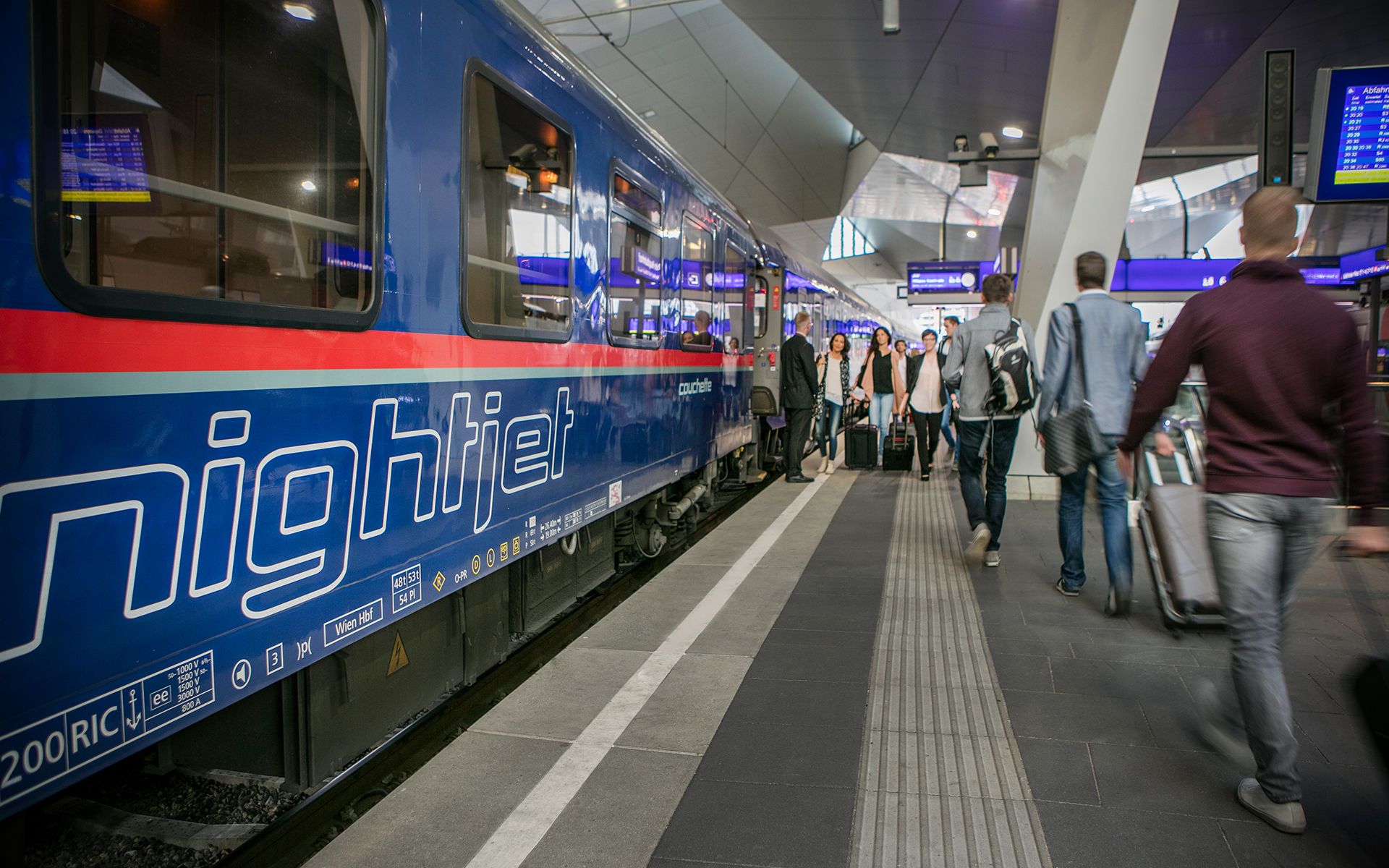 ÖBB Nightjet Schlafwagen am Bahnsteig eines modernen Bahnhofs. Der blaue Zug mit weißer Nightjet-Beschriftung und rotem Streifen steht bereit. Fahrgäste mit Gepäck bewegen sich auf dem beleuchteten Bahnsteig.