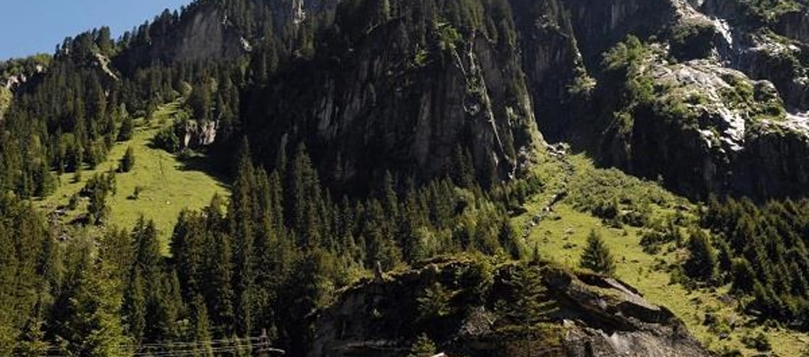 A cabin at the base of a lush, forested mountain with rocky cliffs and a blue sky above.