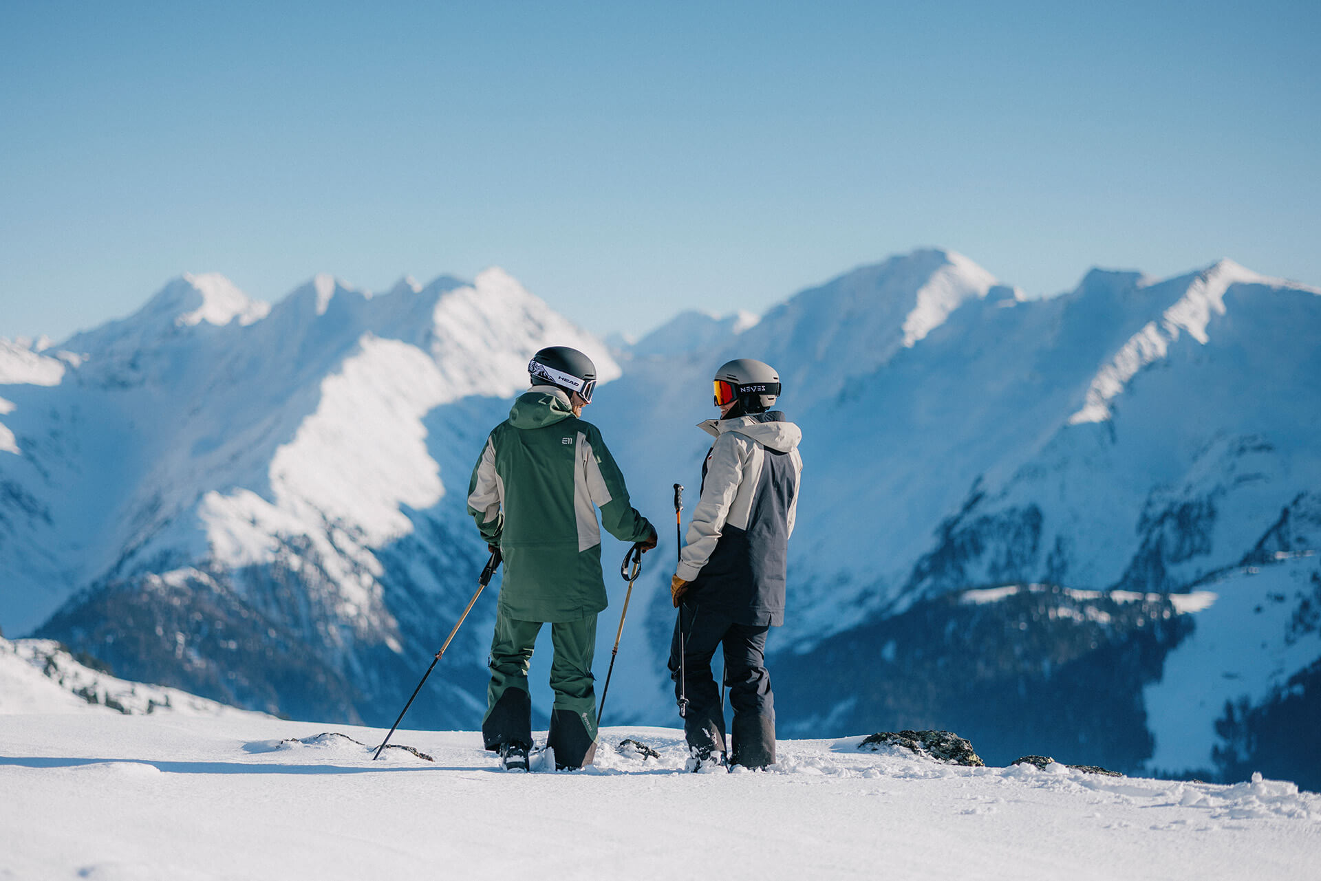 Zwei Skifahrer in Schutzkleidung fahren auf einer verschneiten Piste in den Bergen bei Sonnenschein.