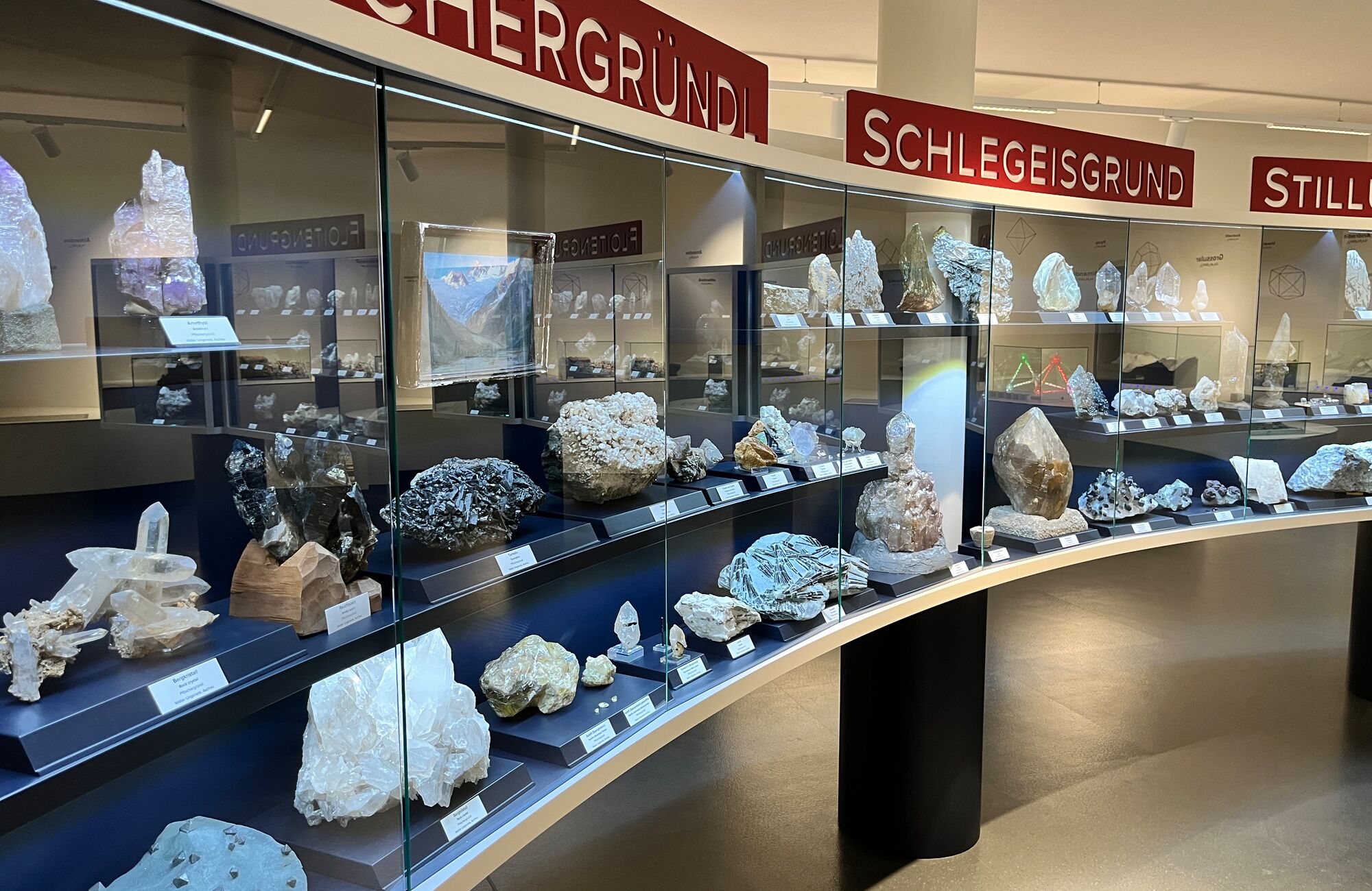 Exhibition at the Nature Park House Ginzling in the Zillertal with numerous minerals and rock formations displayed in illuminated showcases. The red signs above the showcases indicate different categories of exhibits.