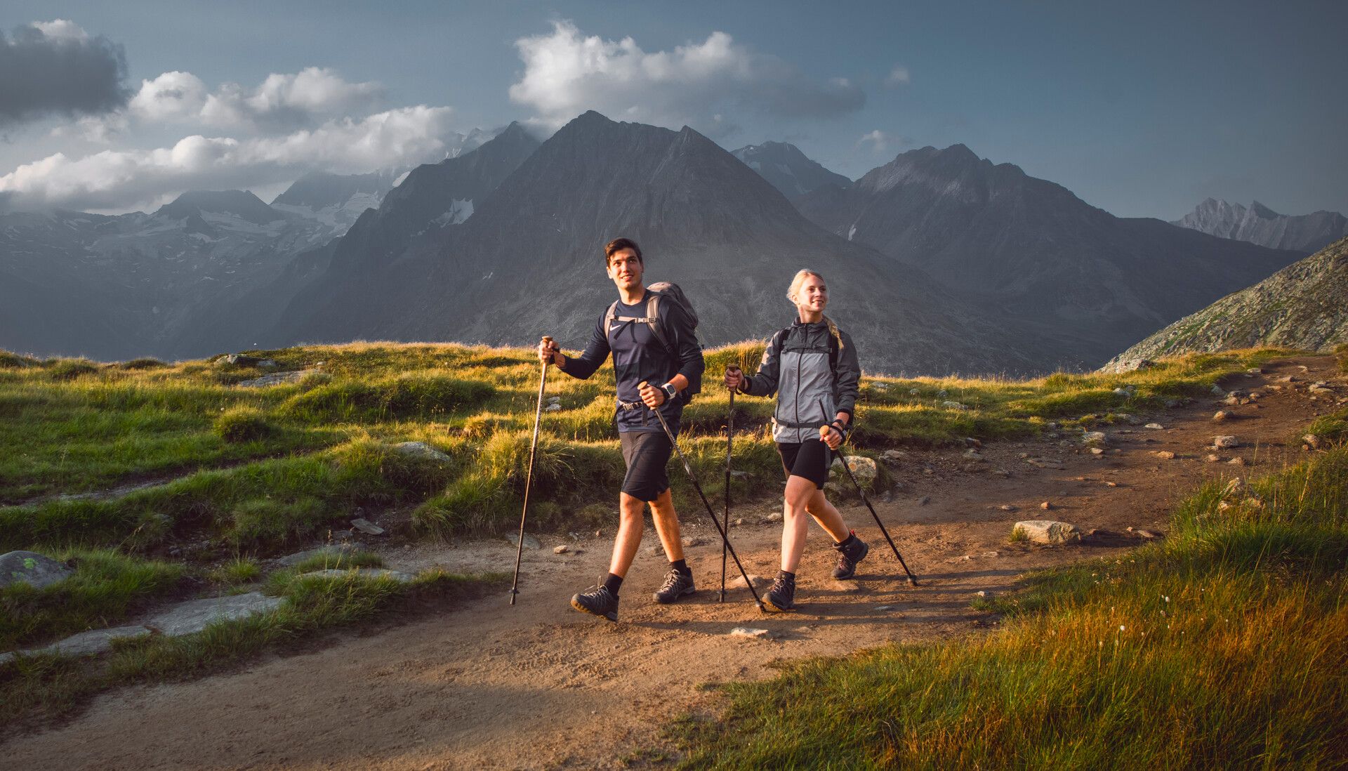 Two people hike with poles on a mountain trail. Around them are green meadows and small streams. In the background, tall snow-covered mountains under a cloudy sky. Sunlight casts a warm, natural glow on the scene.