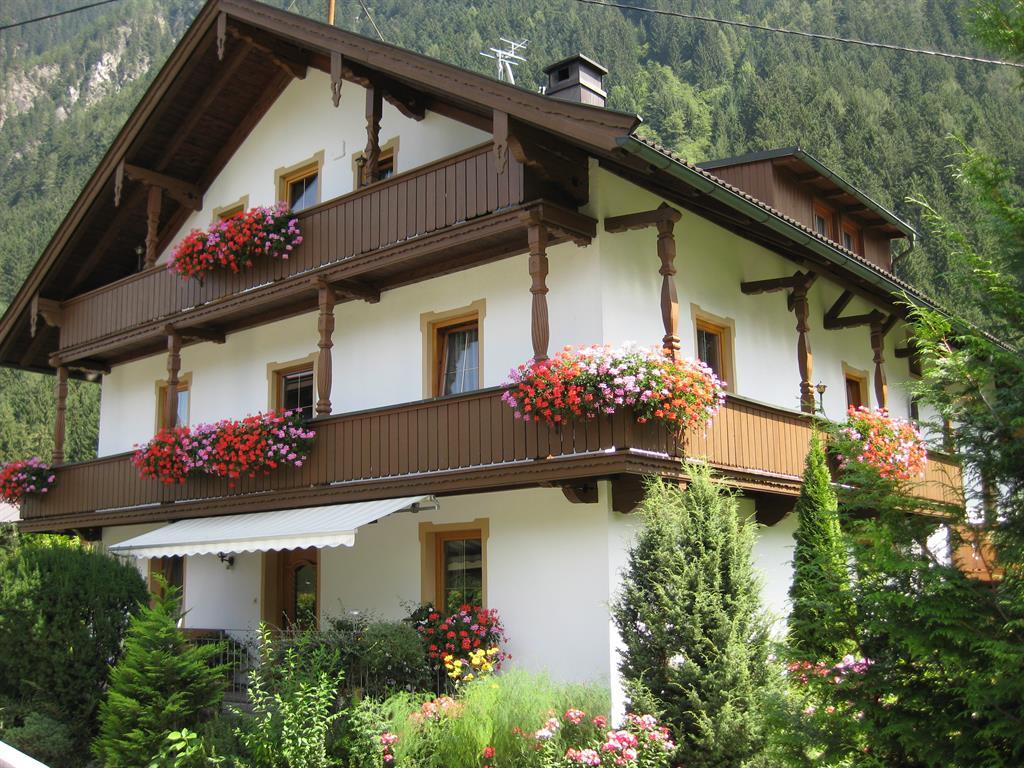 House with white walls, wooden balconies, and colorful flowers in front of green mountains.