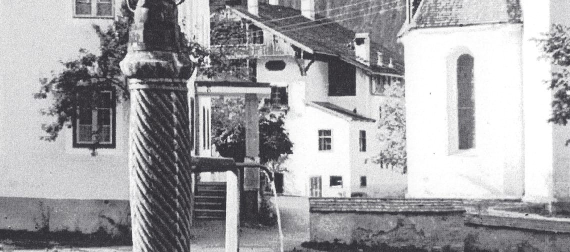 Black-and-white photo from 1928: Marienbrunnen in Mayrhofen – fountain with a column and basin in the foreground; the parish church and the Widum (rectory) behind, with houses and a mountain slope.