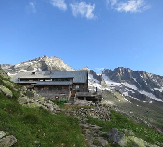 Cabin on a hillside with green grass, mountains with snow patches in the background, and a clear blue sky.