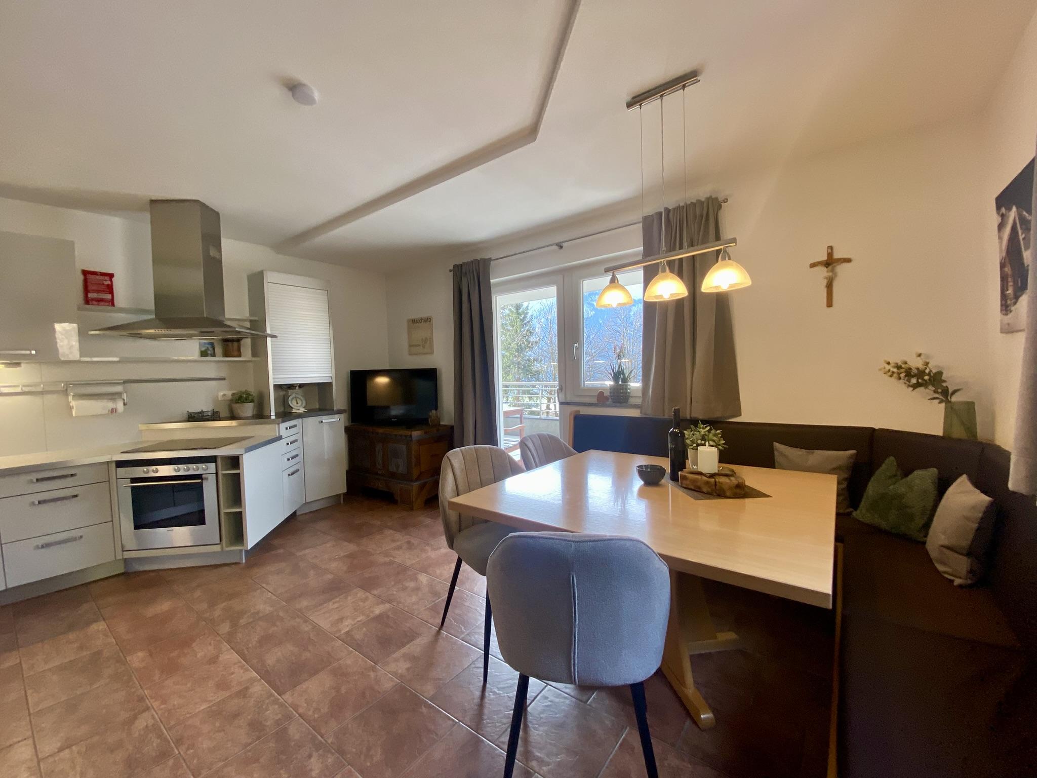 Bright living area with dining table, chairs, kitchen, window, and wall decorations including a cross.
