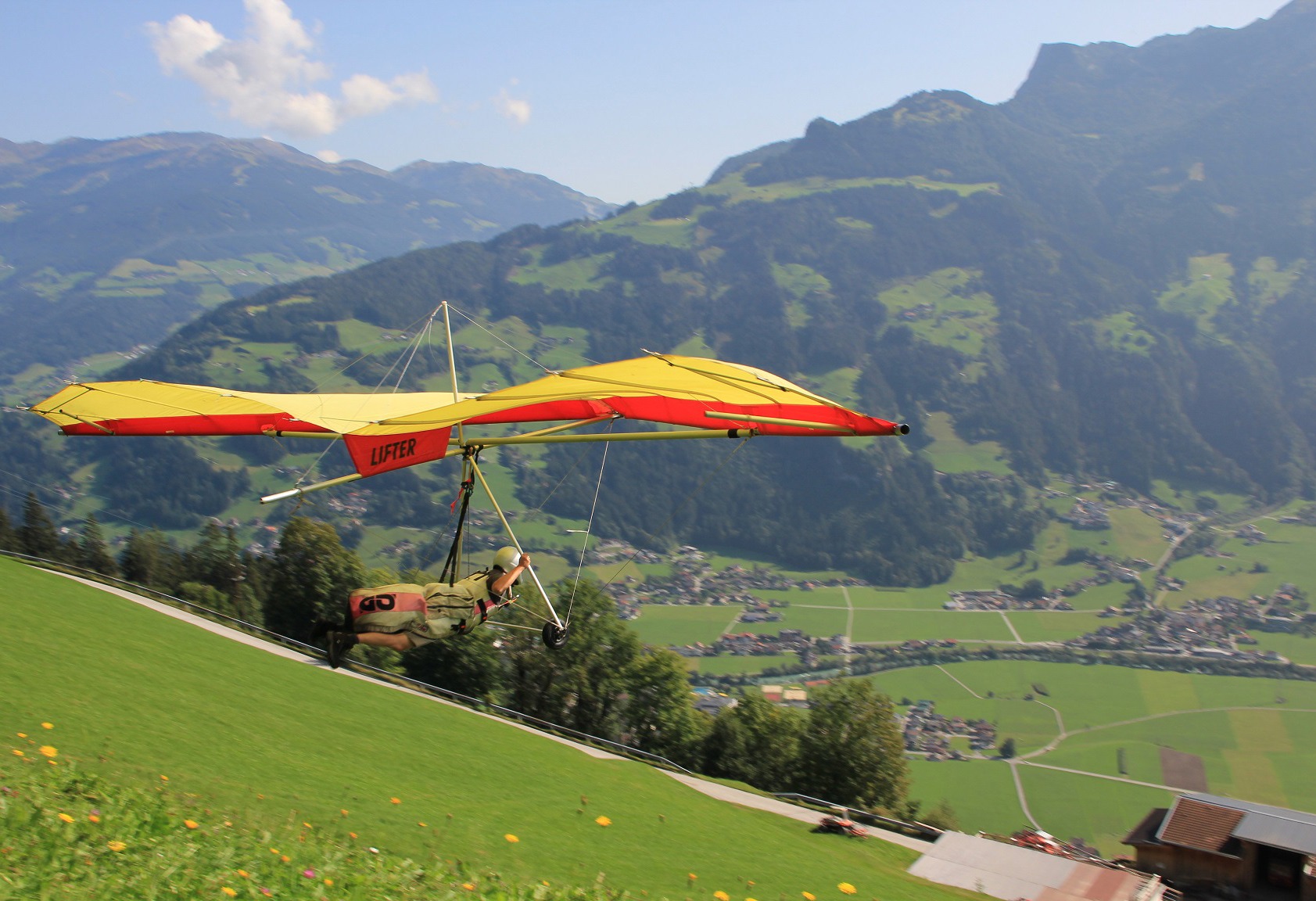 A hang glider pilot takes off from a green mountain meadow in Mayrhofen, Tyrol, with a red-yellow glider. The Zillertal valley with mountains and villages is in the background. Perfect flying day with a blue sky.