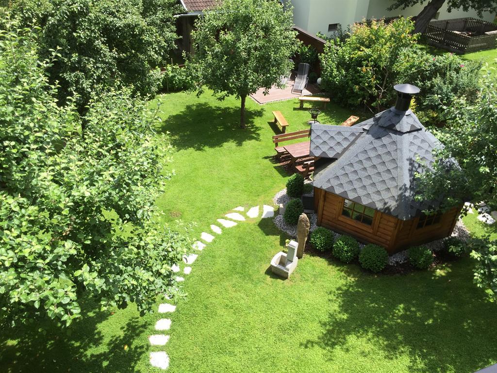 Garten mit Baum, Steinen, Holzhaus, Bänken und Pflanzen im Sonnenschein, Blick von oben.