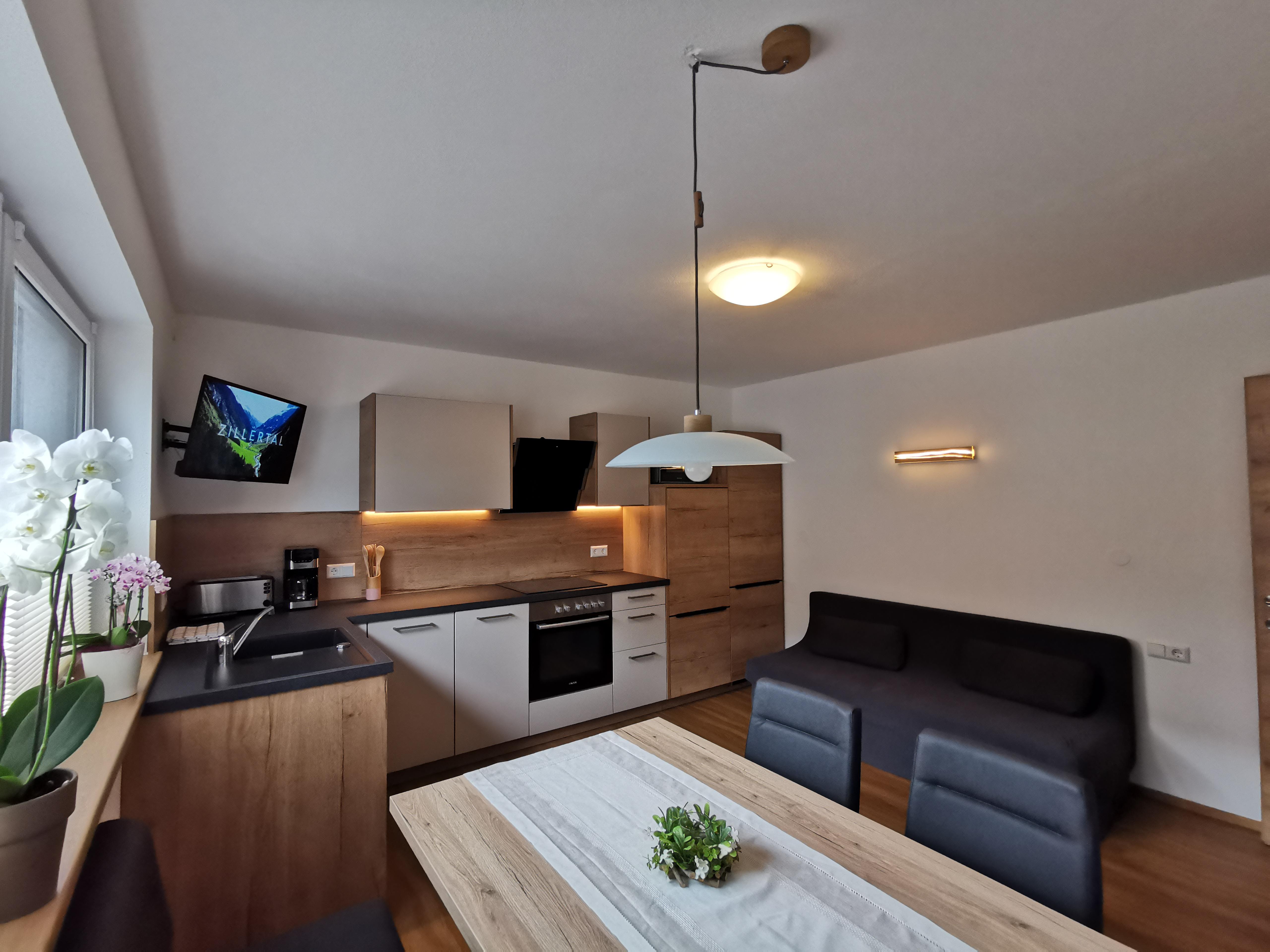 Moderne Küche mit weißen und Holzschränken, Esstisch, Pflanzen und Fernseher an der Wand.