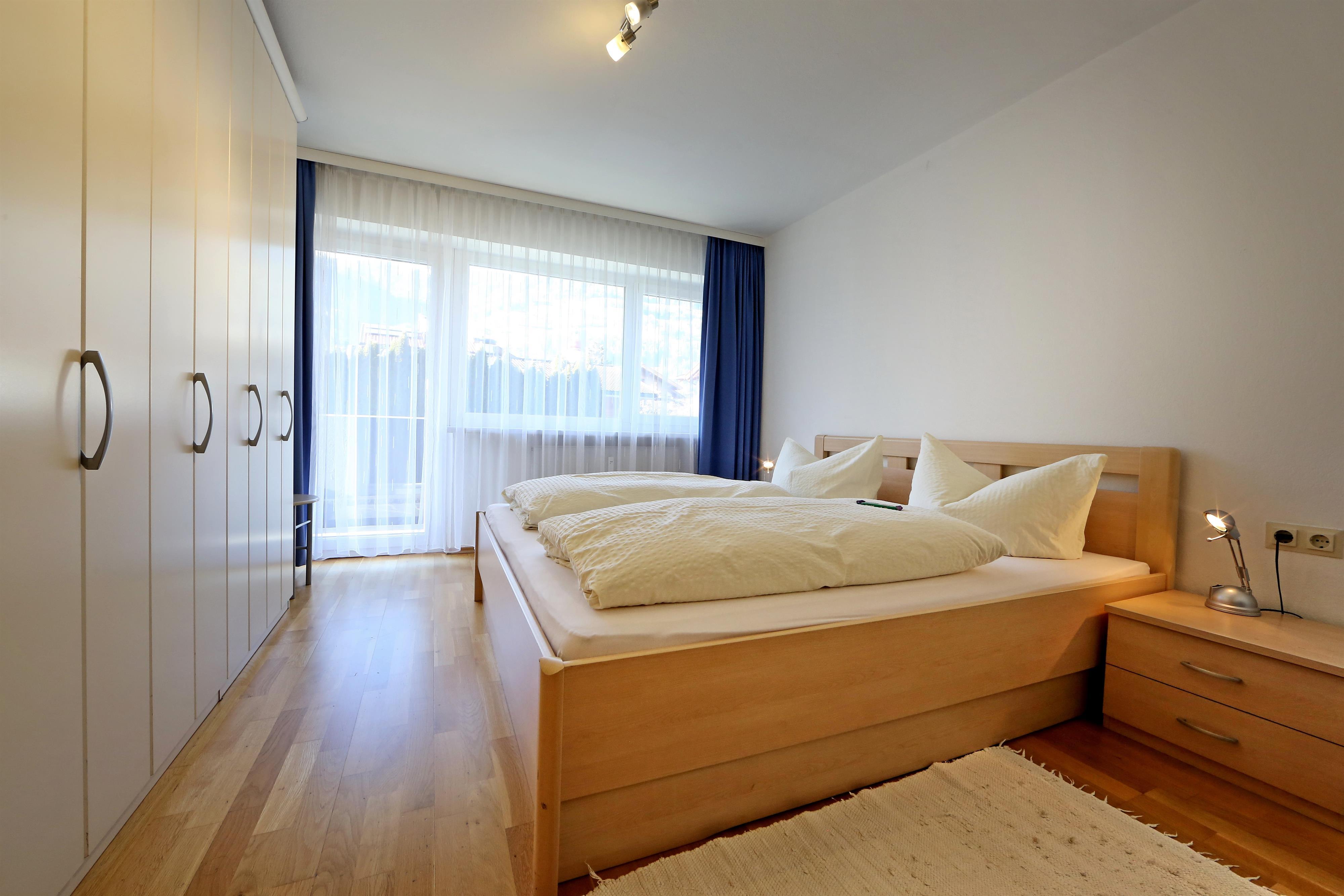 Schlafzimmer mit Doppelbett, Nachttisch, Kleiderschrank, große Fenster mit Vorhängen, Holzfußboden.