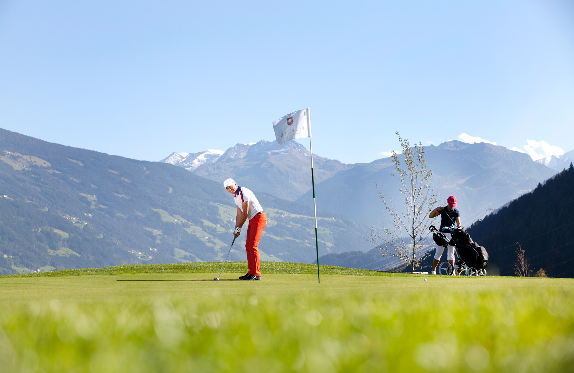 Two people golfing at Uderns Golf Course in Zillertal. One is teeing off, the other stands with a golf bag. Snowy Alps in the background – golf and nature experience in Tyrol.