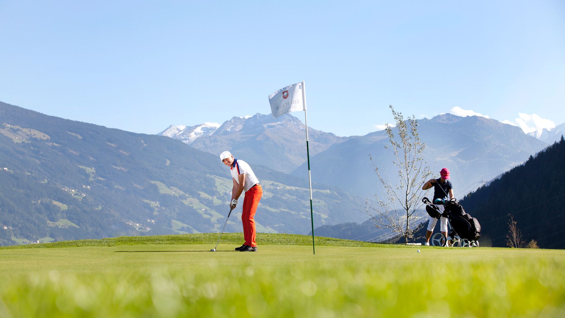 Two people golfing at Uderns Golf Course in Zillertal. One is teeing off, the other stands with a golf bag. Snowy Alps in the background – golf and nature experience in Tyrol.