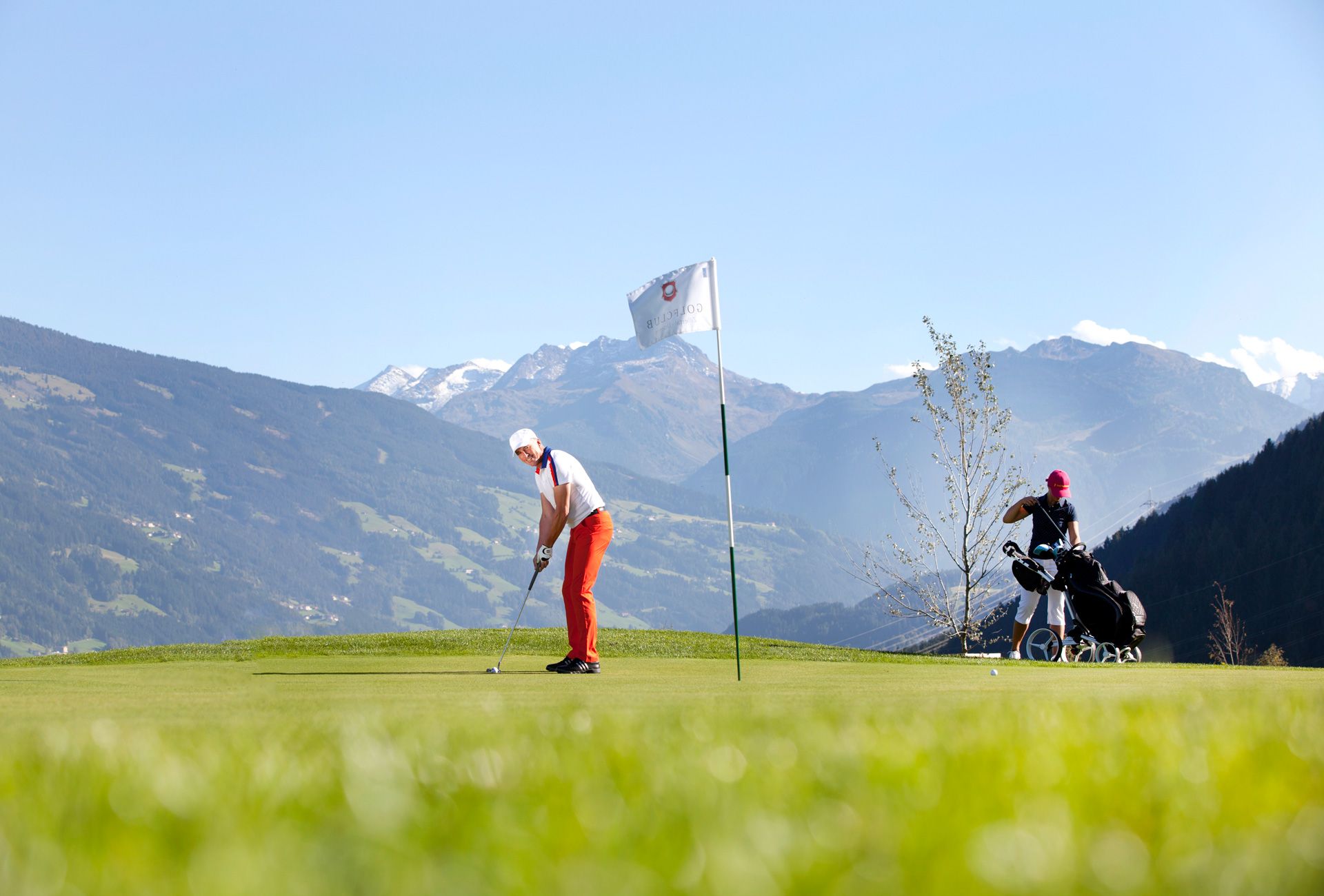 Zwei Personen beim Golfen auf dem Golfplatz Uderns im Zillertal. Eine schlägt ab, die andere steht mit Golfbag. Im Hintergrund die schneebedeckten Zillertaler Alpen – Golf- und Naturerlebnis in Tirol.