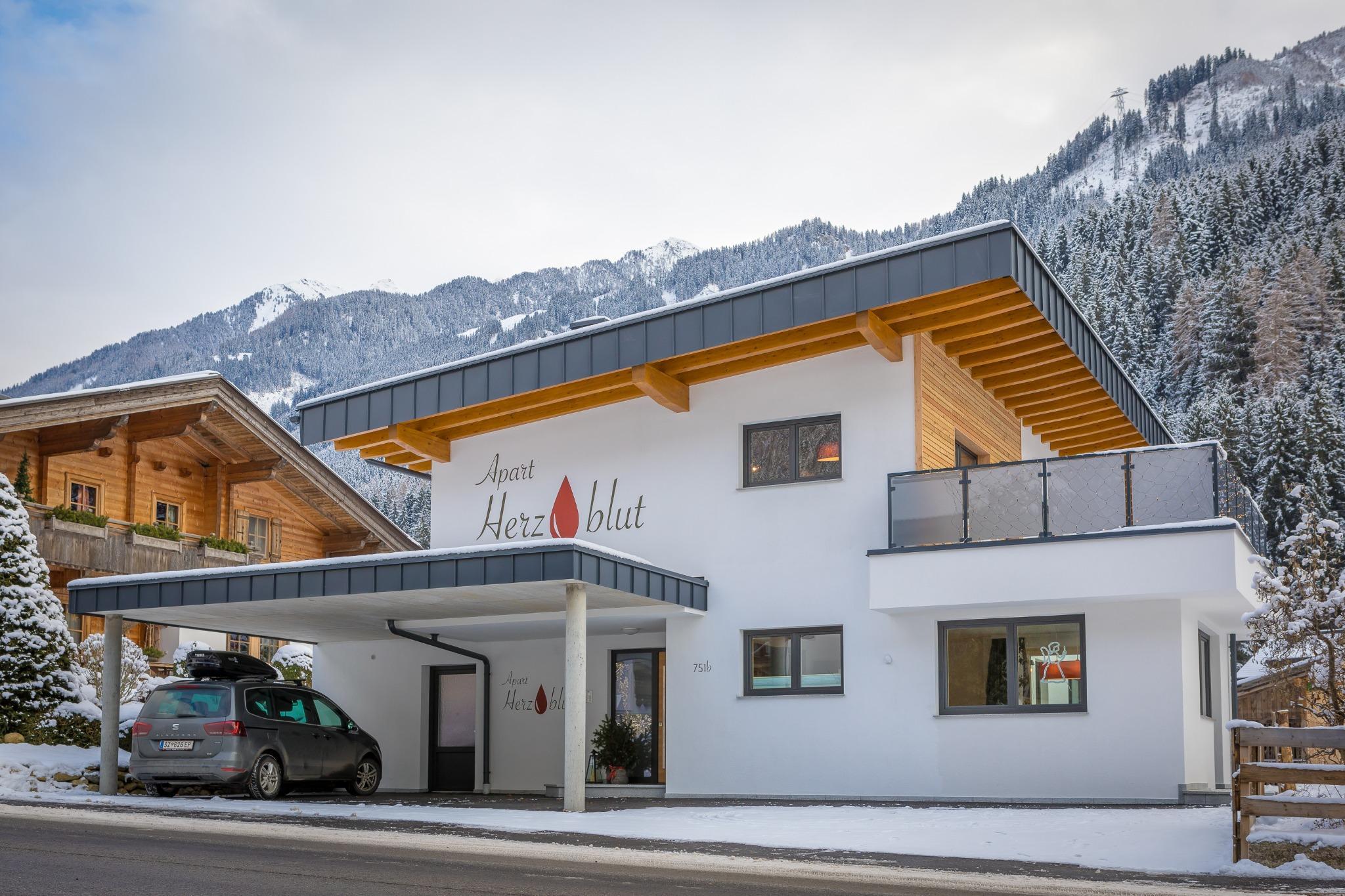 Modern white building with wooden accents and balcony, snowy mountains in the background, car and street in front.