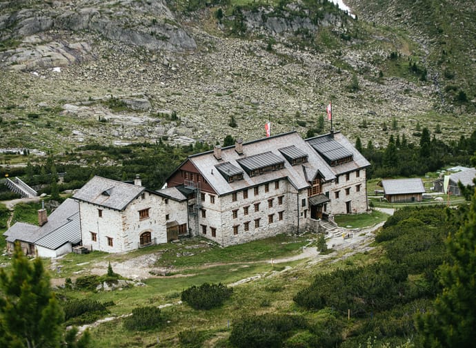 A historic multi-story house in the mountains with stone walls and roof, surrounded by greenery and rocks.