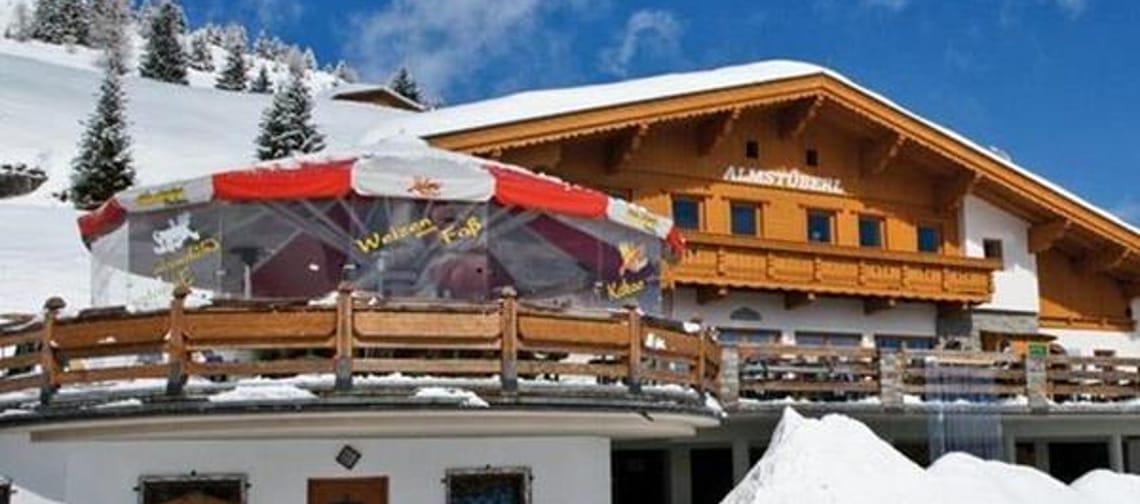 A snow-covered chalet with a large balcony, a red umbrella, and snow-covered trees in the background.