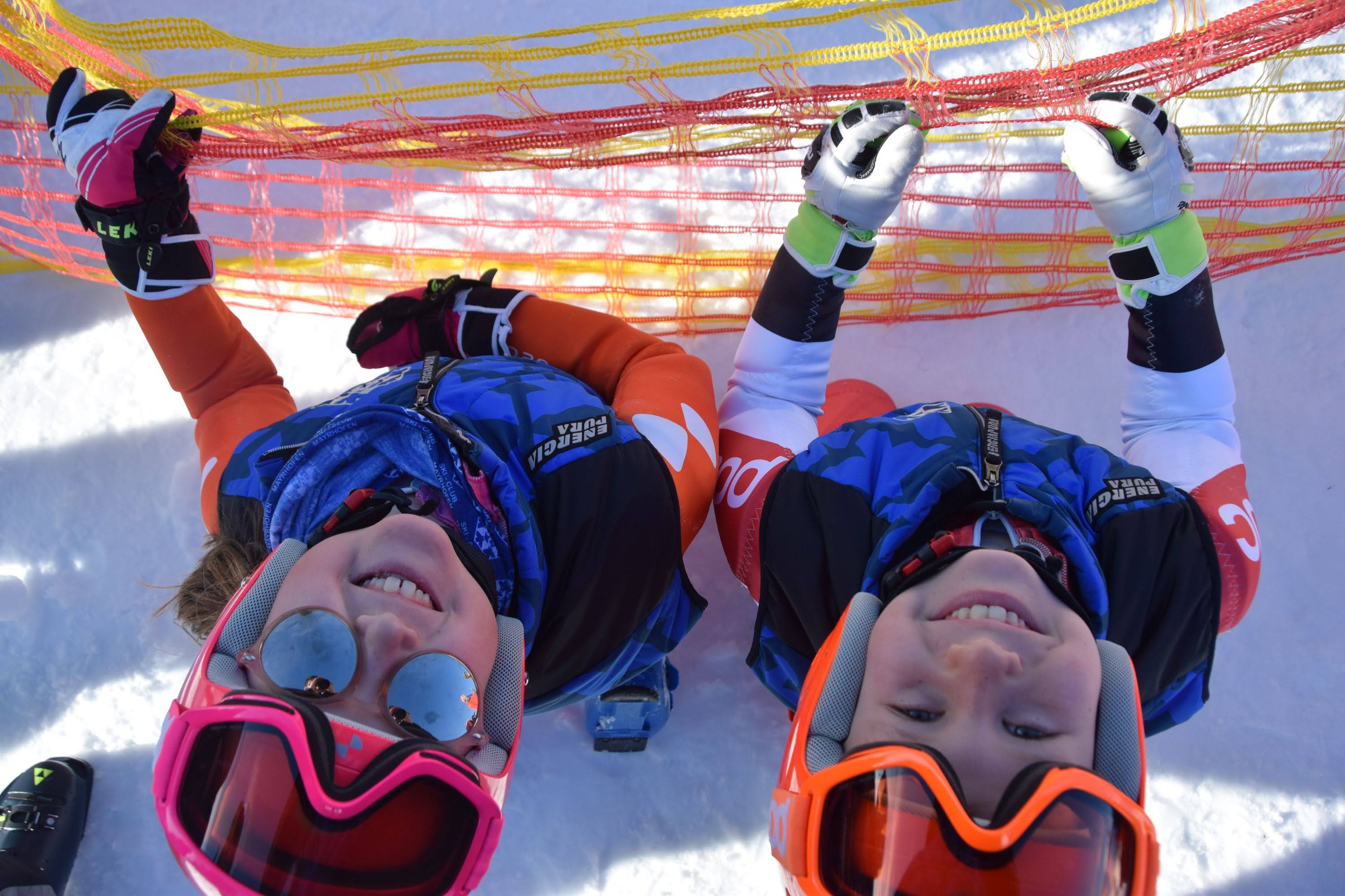 The picture shows two children in colorful ski clothing, laughing under a safety net and looking up. They are wearing ski goggles and helmets, and the sunny day and the snow around them convey a cheerful winter sports atmosphere.