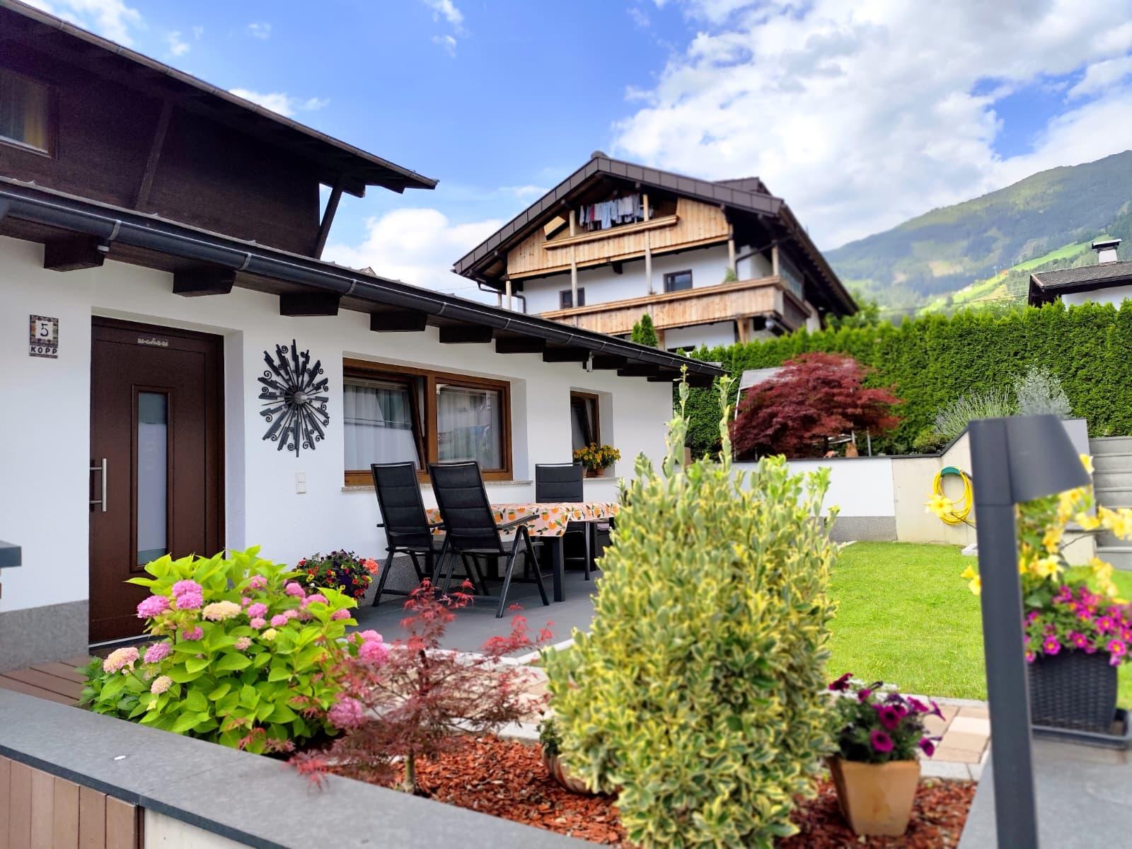 Garten mit blühenden Pflanzen, Terrasse und Blick auf Häuser und Berge im Hintergrund.