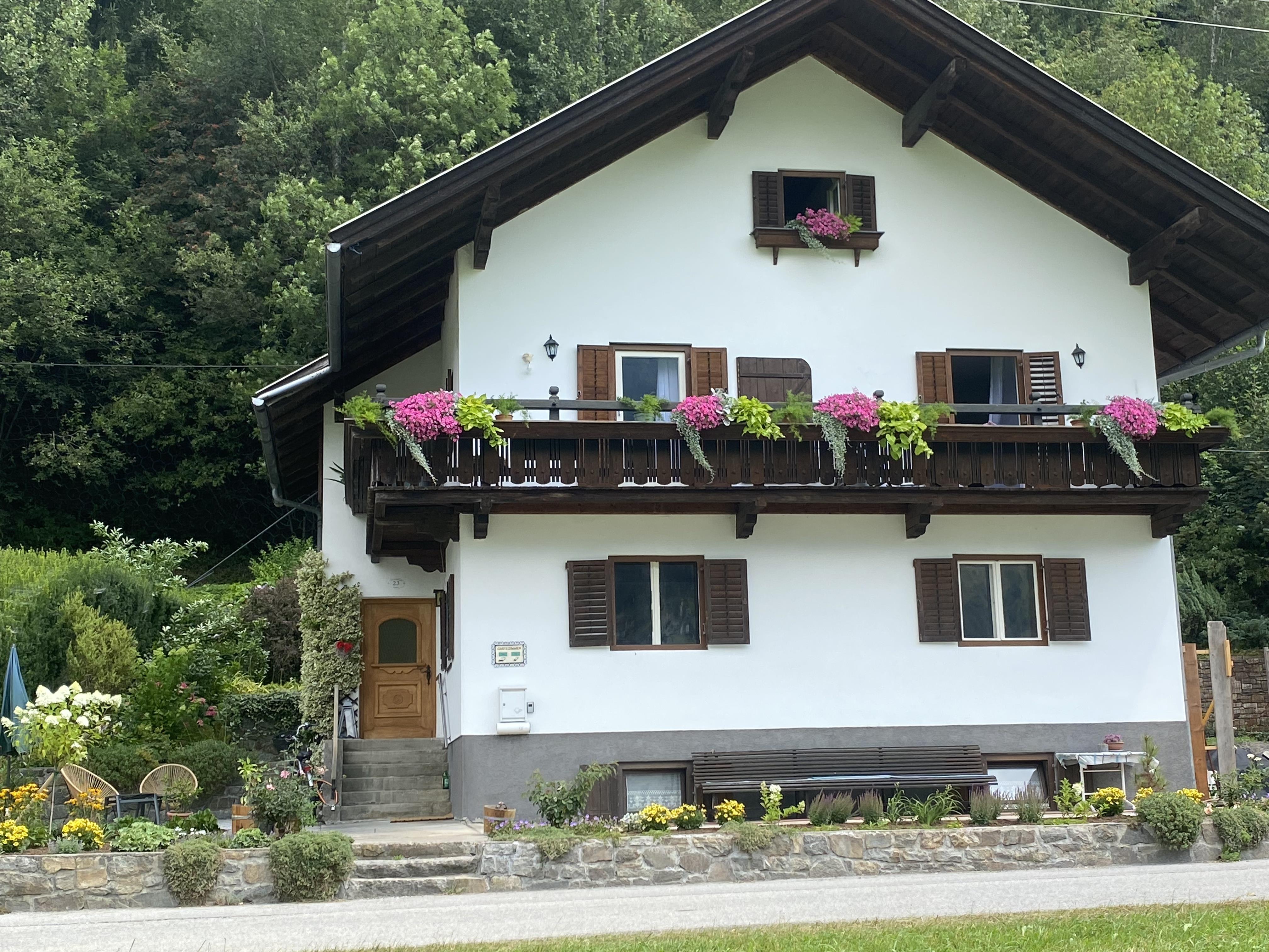 Häuserfassade mit Balkon, blühende Blumen und grünem Garten vor einem bewaldeten Hügel.