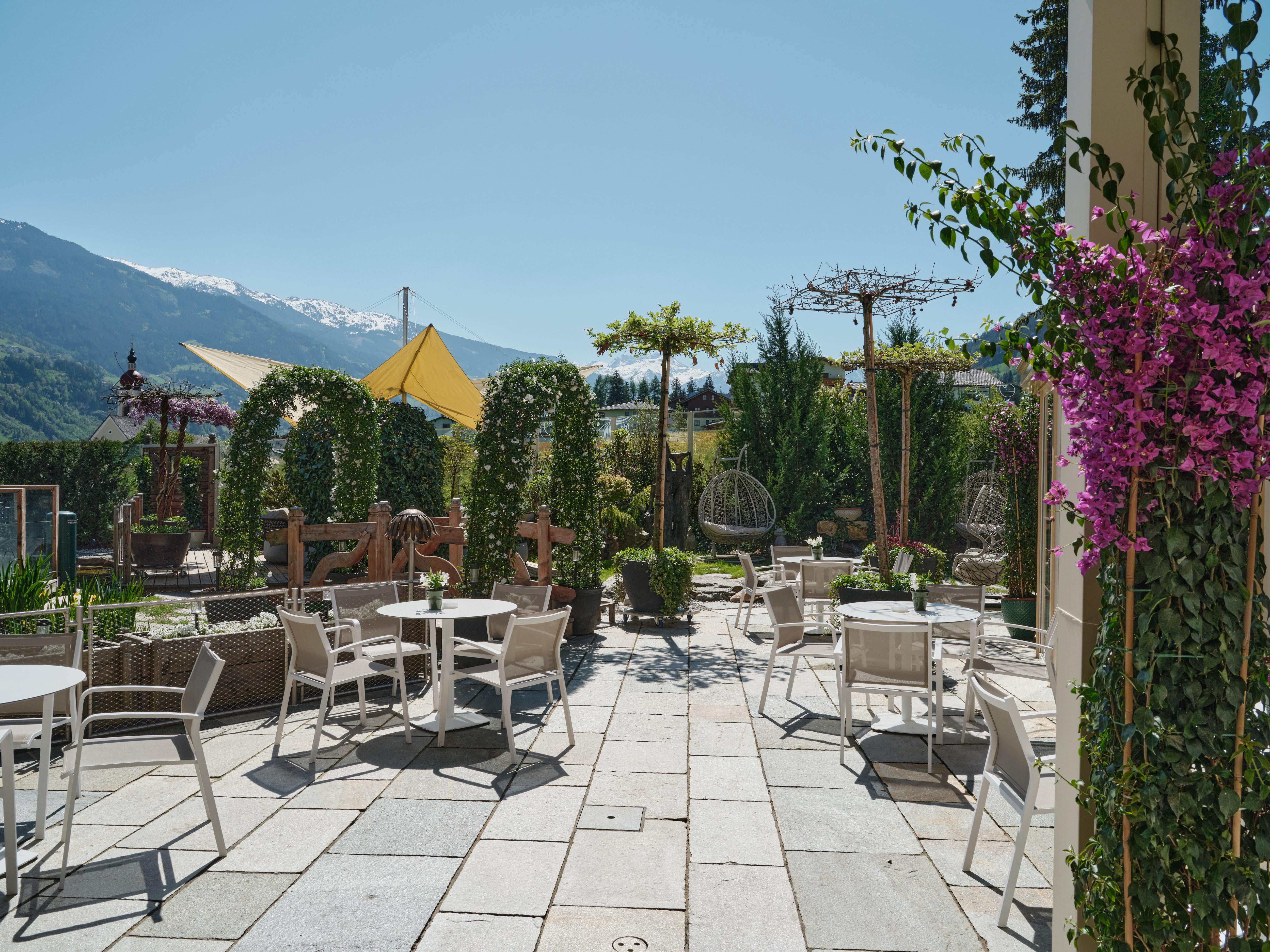 Ein sonniger Garten mit weißen Tischen, Stühlen, blühenden Pflanzen und Bergen im Hintergrund.