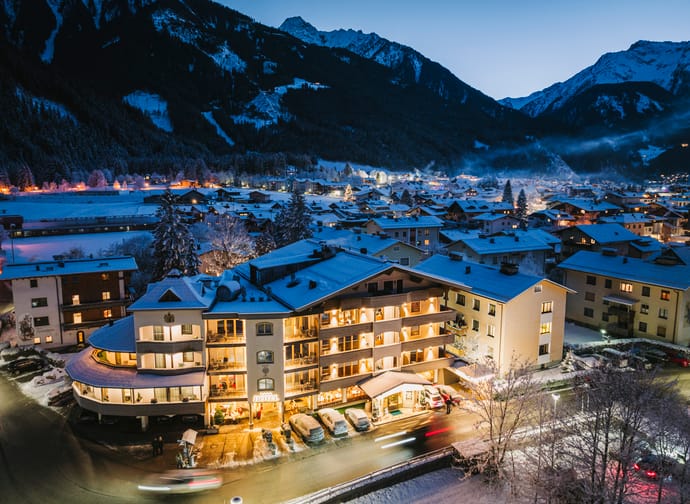 Mehrstöckiges Hotel in verschneitem Bergdorf bei Nacht, mit beleuchteten Fenstern und umliegenden Bergen.