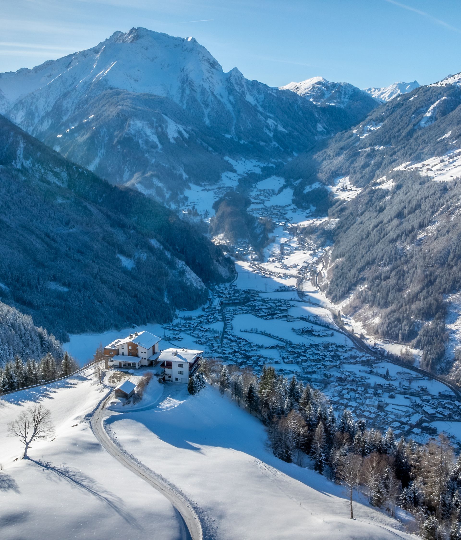 Winterpanorama Brandberg im Zillertal: Luftaufnahme des verschneiten Bergdorfs auf sonnigem Südhang. Traditionelle Bauernhöfe zwischen weißen Wiesen, umgeben von bewaldeten Bergen. Im Hintergrund das Zillertal mit schneebedeckten Gipfeln.