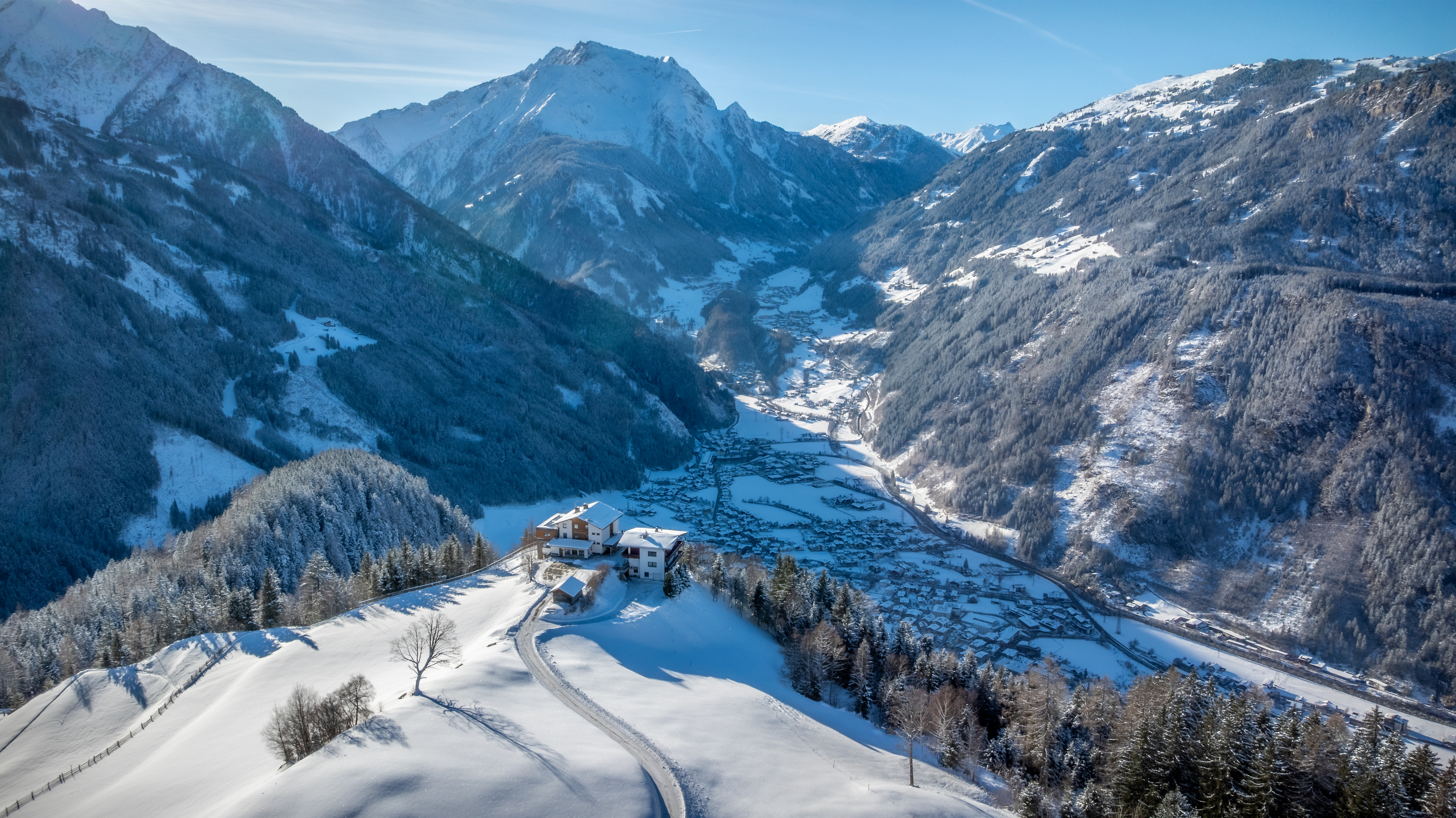 Winter panorama of Brandberg in Zillertal: Aerial view of the snow-covered mountain village on a sunny south-facing slope. Traditional farmhouses among white meadows, surrounded by forested mountains. Zillertal valley with snow-capped peaks in background.