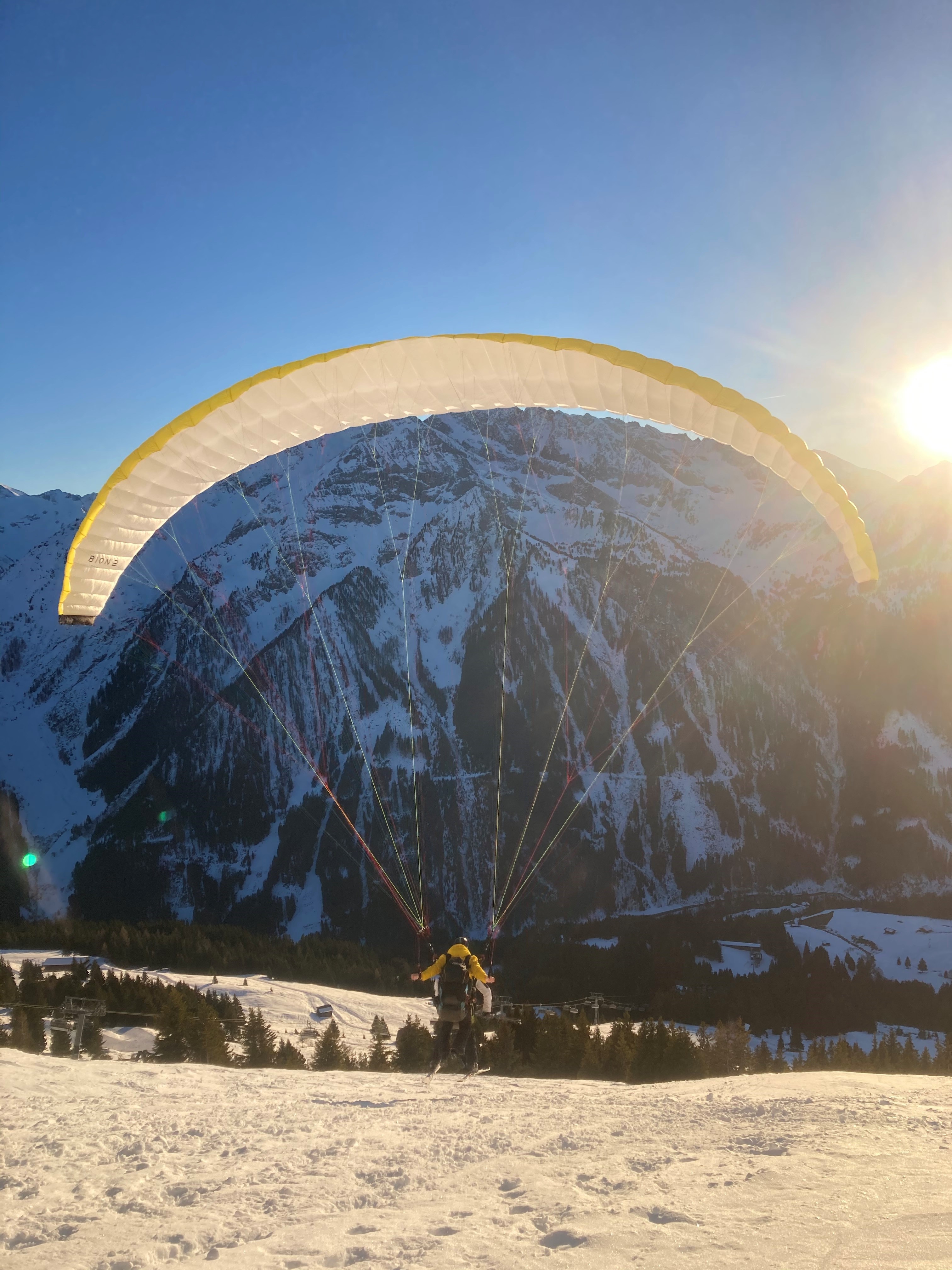 Person paragliding over snowy mountains at sunset, with a clear blue sky in the background.