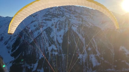 Person beim Paragliding in verschneiter Berglandschaft bei Sonnenuntergang, mit blauer Himmel im Hintergrund.