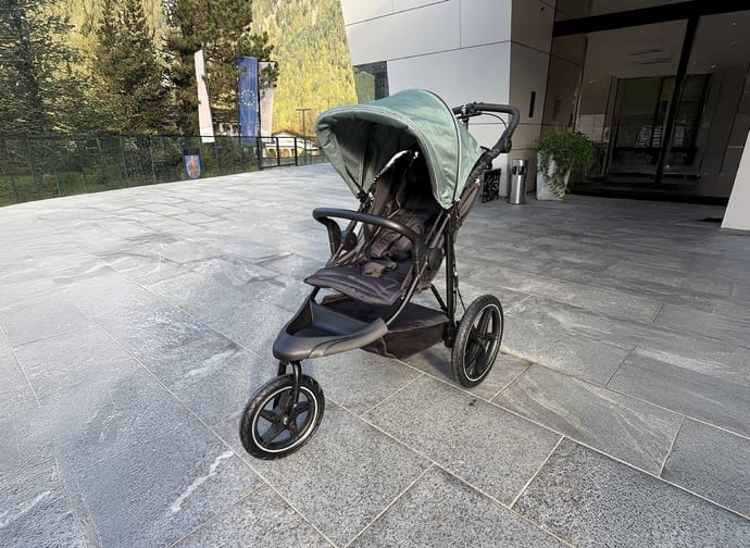 Ein schwarzer Kinderwagen mit grünem Verdeck steht auf einem breiten, glatten Gehweg vor einem modernen Gebäude in Mayrhofen. Der Zugang ist ebenerdig und gut befahrbar.