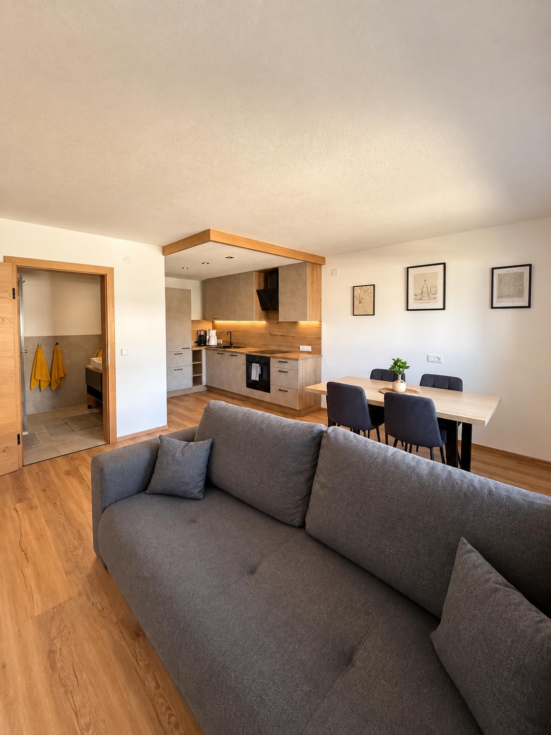 Moderner Wohnraum mit grauem Sofa, offener Küche mit Holzakzenten und Esstisch mit vier Stühlen.
