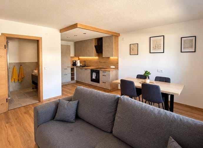 Moderner Wohnraum mit grauem Sofa, offener Küche mit Holzakzenten und Esstisch mit vier Stühlen.