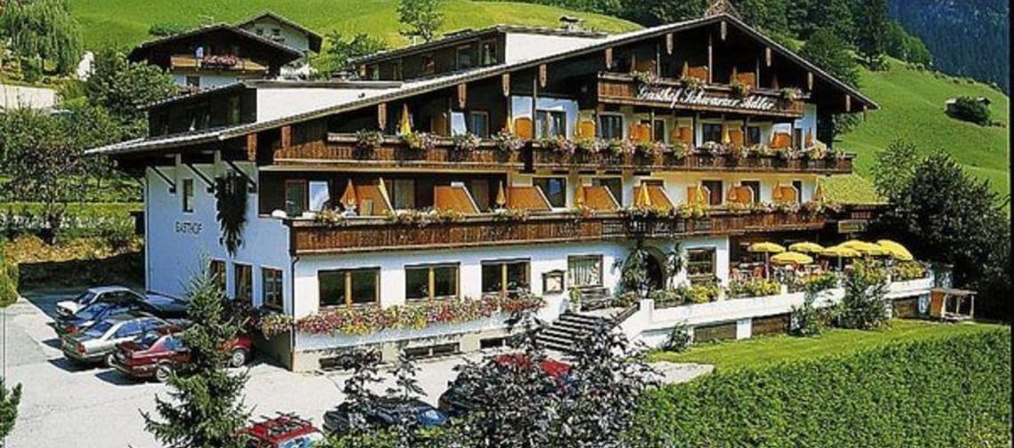 Das Bild zeigt den Gasthof Schwarzer Adler von außen. Vor dem Gebäude erstreckt sich ein offenes Feld, und auf der Terrasse stehen mehrere gelbe Sonnenschirme, die für eine einladende Atmosphäre sorgen.