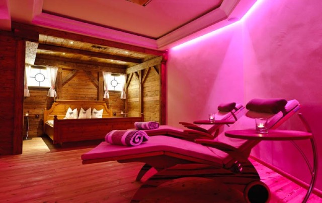 Das Bild zeigt mehrere Massageliegen, neben denen kleine Tische stehen. Dahinter befindet sich ein Holzbett, das von einem sanften pinken LED-Licht beleuchtet wird, was eine entspannende Atmosphäre schafft.