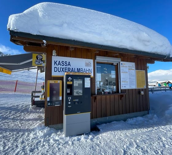 Kassenhäuschen im Schnee mit Ticketautomat, Schild 'Kassa Duxeralmbahn' und Skilift im Hintergrund.