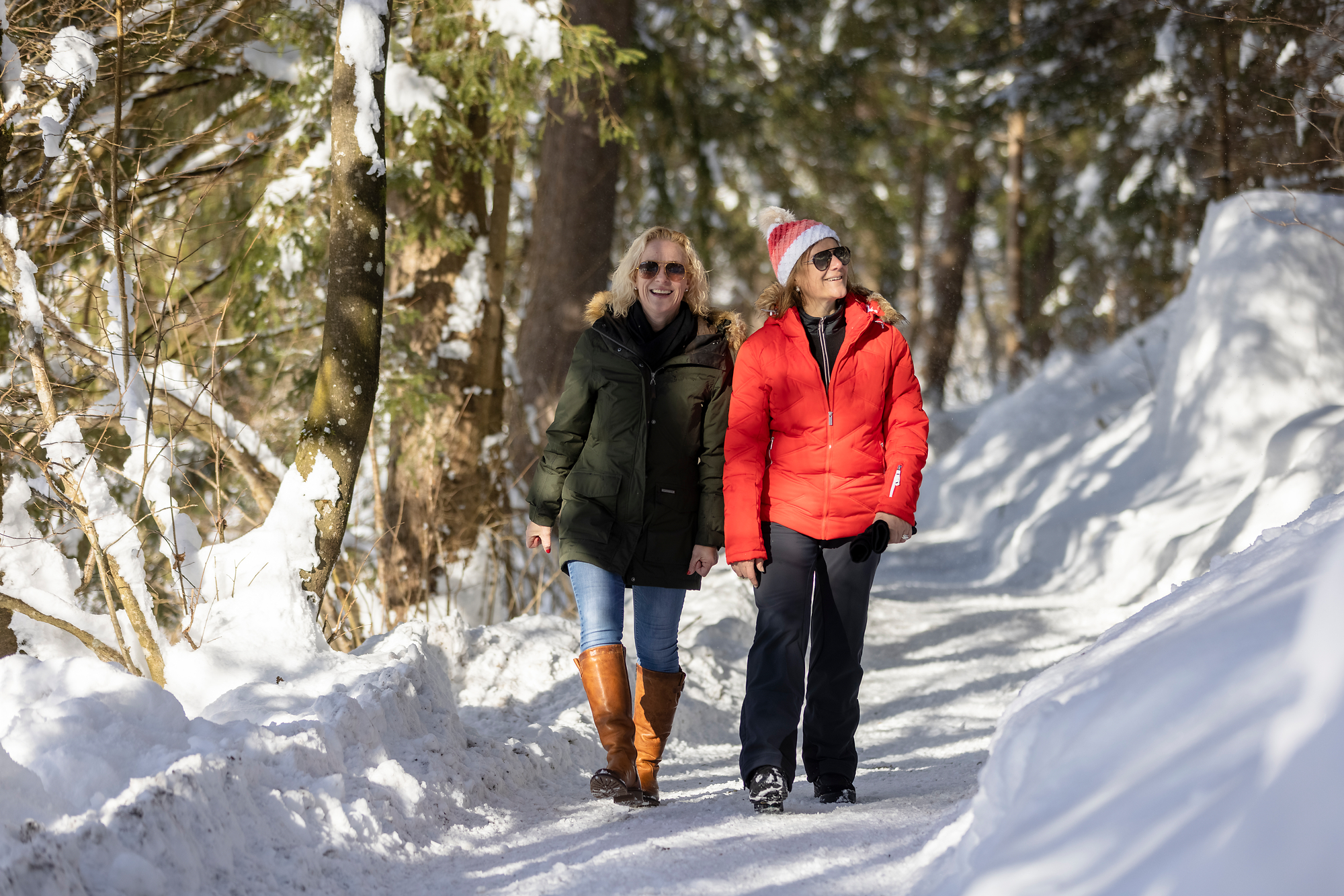 Zwei Frauen beim Winterwandern auf einem verschneiten Waldweg in Mayrhofen-Hippach, Tirol. Sie tragen warme Winterkleidung und genießen bei Sonnenschein die verschneite Natur.