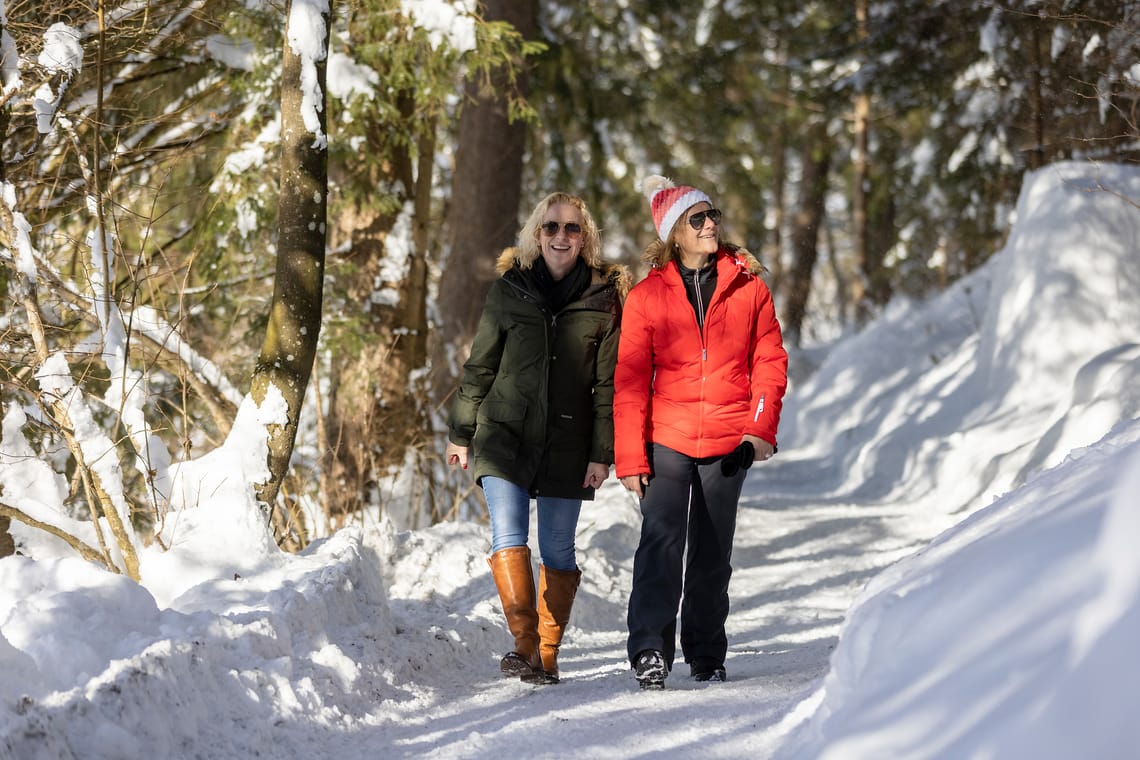 Wandern am Easy Trail in Mayrhofen-Hippach im Winter ©Thomas Eberharter Photography Zwei Frauen beim Winterwandern auf einem verschneiten Waldweg in Mayrhofen-Hippach, Tirol. Sie tragen warme Winterkleidung und genießen bei Sonnenschein die verschneite Natur.