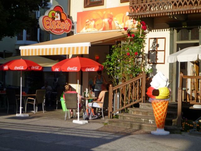 Das Bild zeigt die Gelateria Del Corso von außen. Die Fassade ist weiß, während große orangefarbene Akzente, darunter eine orange Markise, dem Gebäude einen lebendigen Look verleihen. Vor der Tür steht ein großes Modell eines Eises, und das Logo mit einem